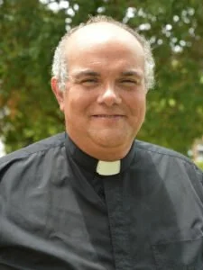 A middle-aged man in clergy attire outdoors, with trees in the background.