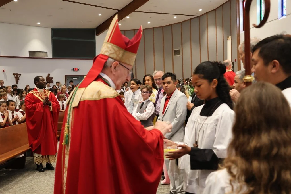 A religious ceremony inside a church with a bishop in red robes and mitre distributing communion to a line of children and adults, with a congregation in the background.