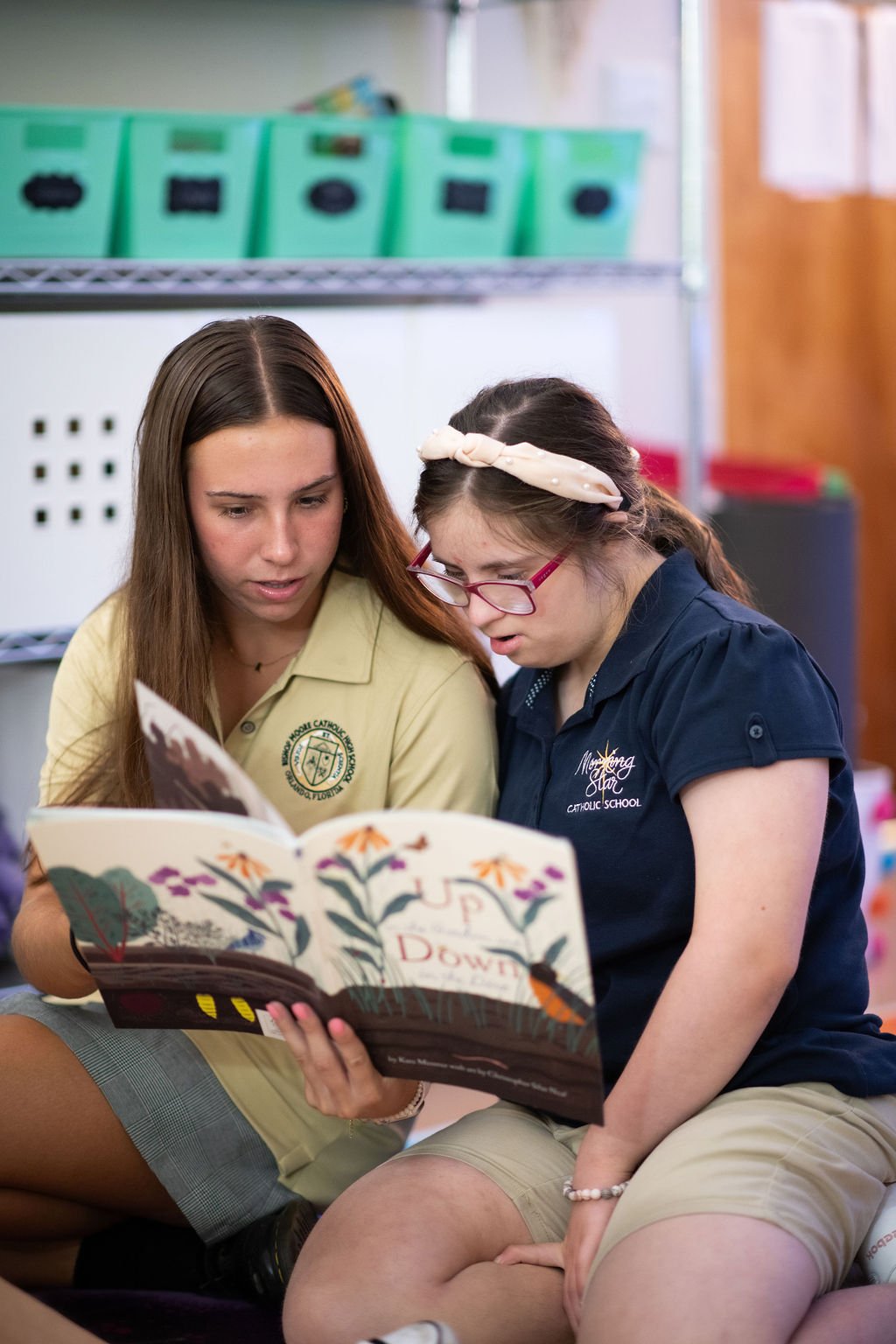Two young girls, one wearing a yellow shirt and the other a navy shirt, sit closely together looking at a colorful book in a classroom.