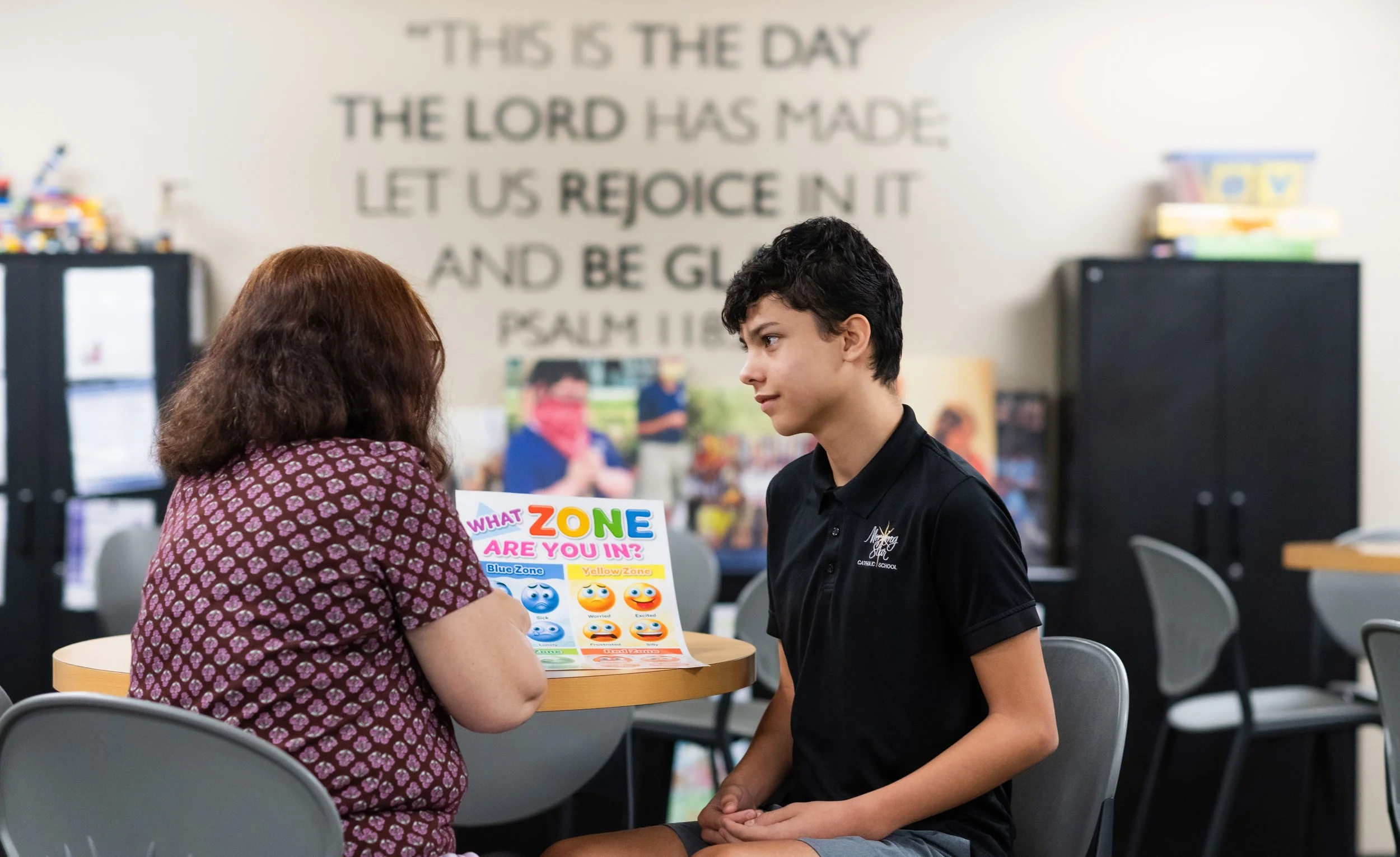 A female instructor with her back to the camera points to a laminated sheet of paper with the emotion zones while young male students looks to her.