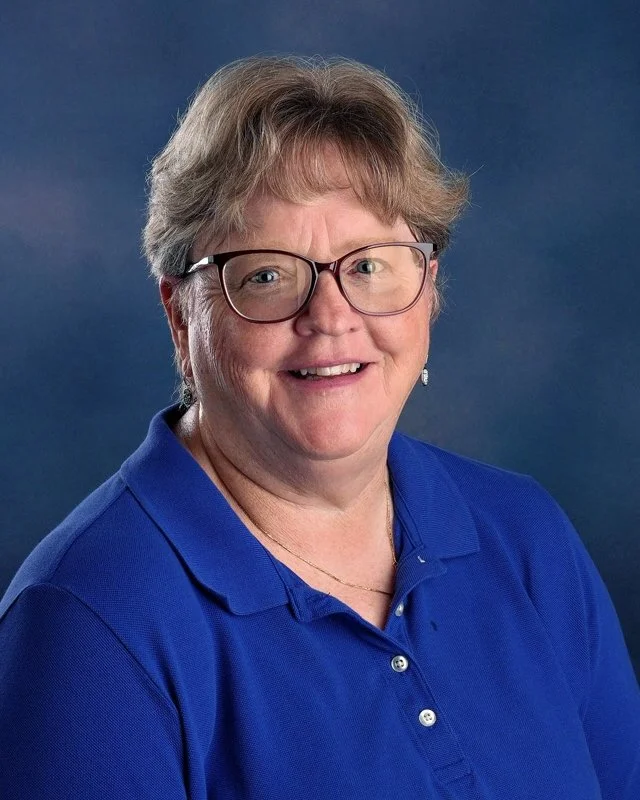 Portrait of a middle-aged woman with short blonde hair, wearing glasses, a blue collared shirt, and a gold necklace, against a blue background.