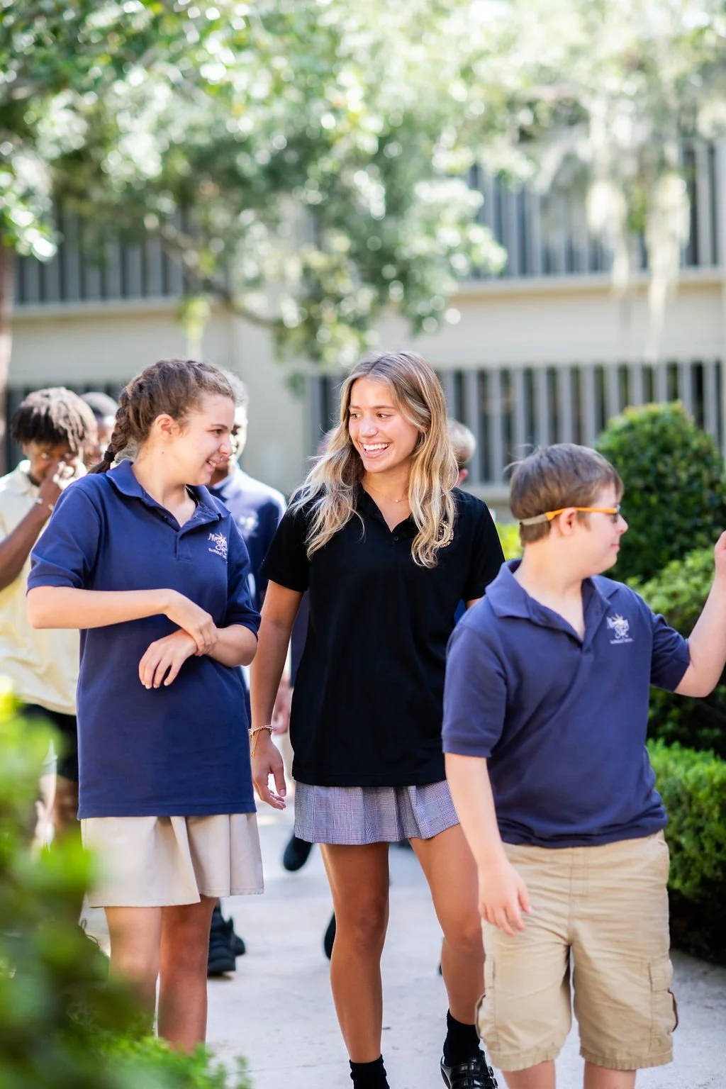 A group of smiling students walking outside on a sunny day, engaging in conversation, with greenery and a school building in the background.
