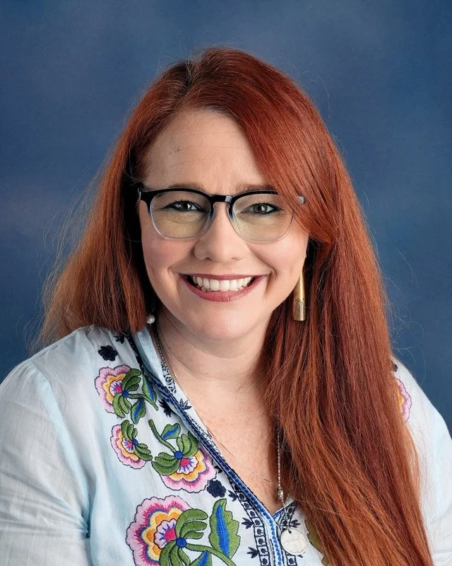 A woman with long red hair, smiling, wearing a black top and gold hoop earrings, against a dark blue background.