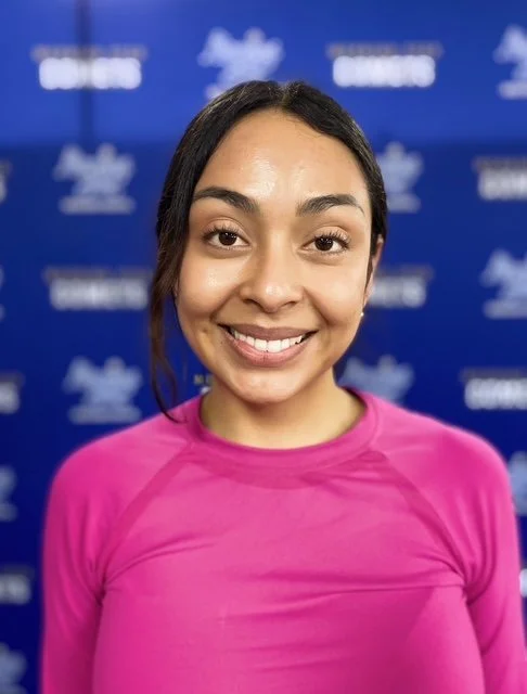 A woman smiling at the camera with shoulder-length gray and light brown hair, wearing a navy blue collared shirt with a necklace, against a blue background.