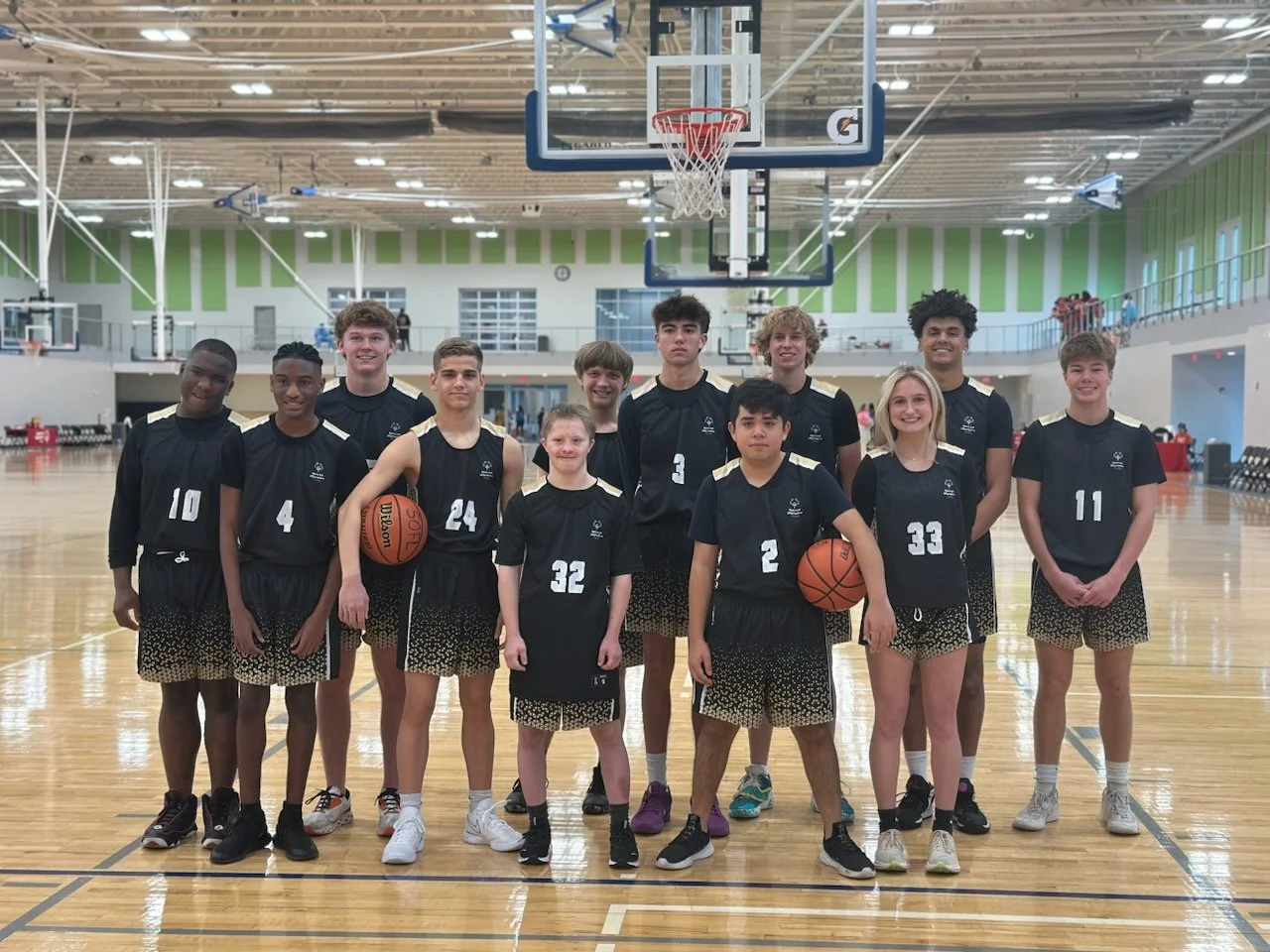 Group of young basketball players posing in a gymnasium with basketball hoops, wearing black jerseys with gold accents and shorts, some holding basketballs.