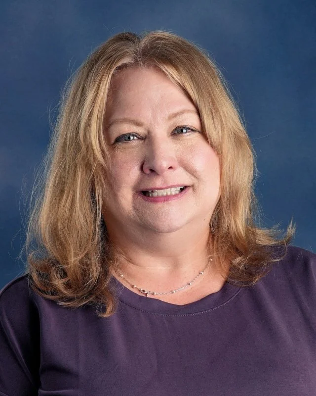 A smiling woman with shoulder-length blonde hair wearing a blue polo shirt and a necklace with a letter 'B'.