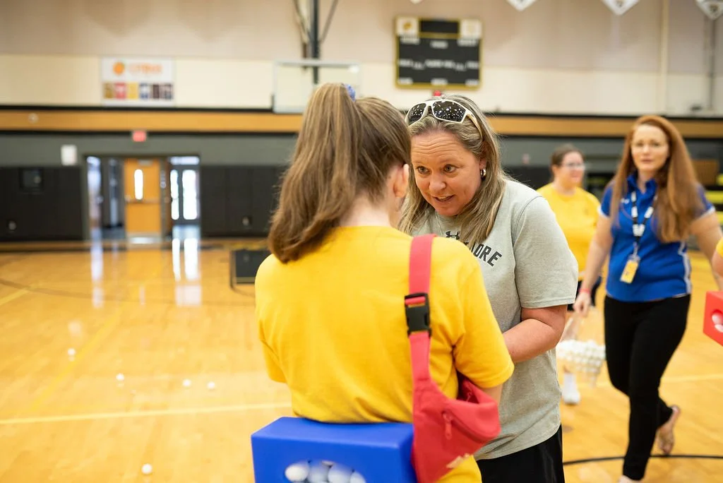 A woman with gray hair and sunglasses on her head talking to a girl in a yellow T-shirt in a gymnasium with a wooden floor, with two other women in the background.