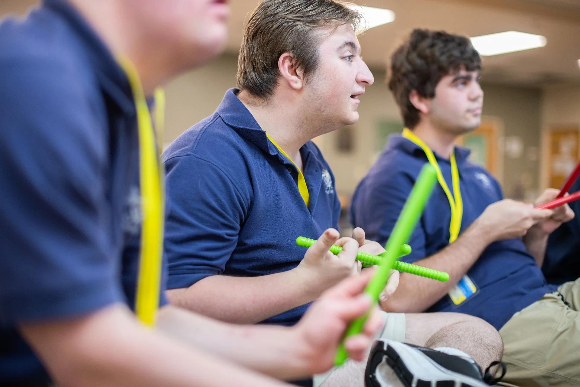 Three young men sitting in a row, holding colorful plastic sticks, in a classroom or indoor setting, wearing blue shirts with yellow accents.