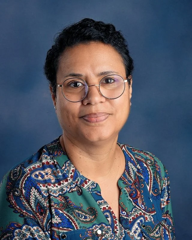 A woman with short curly black hair and glasses, wearing a blue denim shirt, smiling slightly against a blue background.
