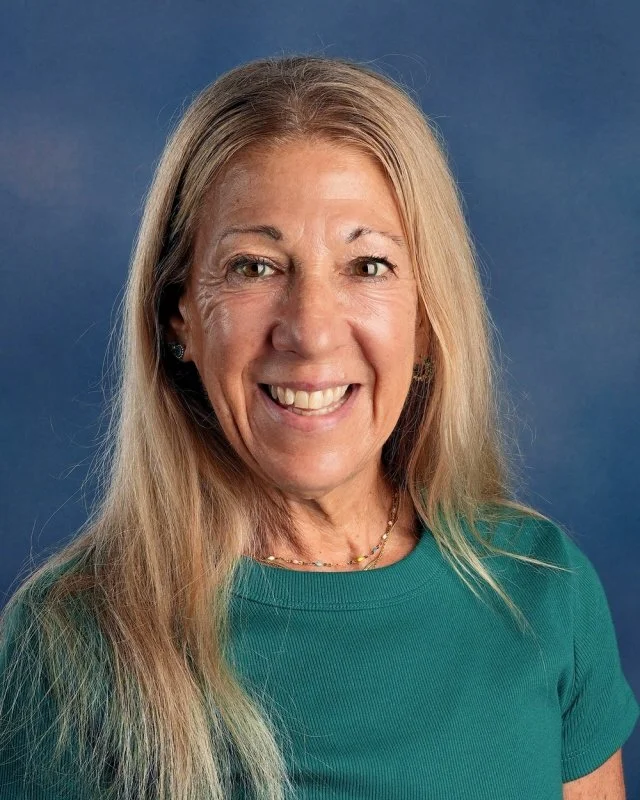 A woman with long blonde hair smiles at the camera against a blue background, wearing a black top and earrings.