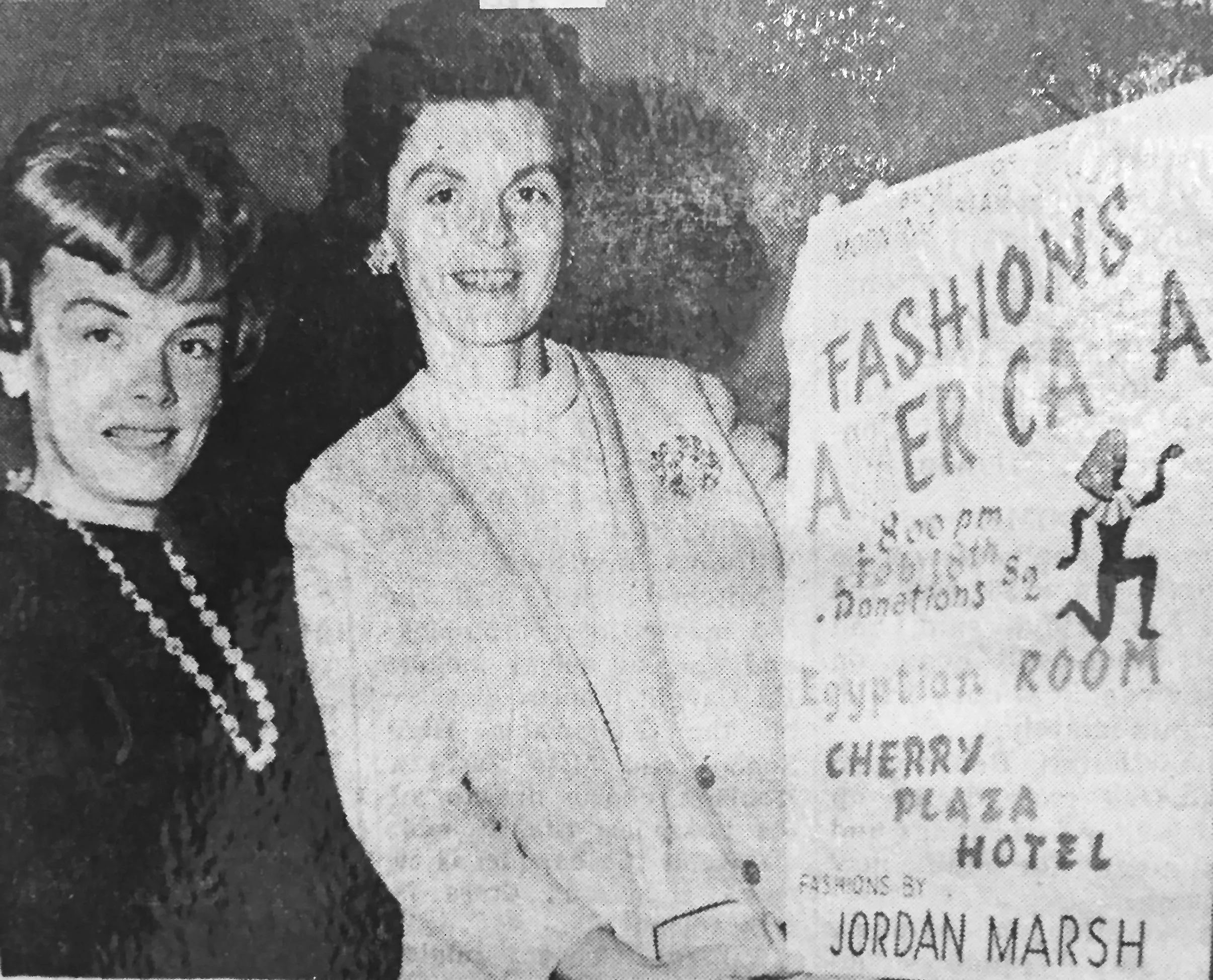 Black and white photo of two women standing next to a sign promoting a fashion show. The woman on the left is wearing a pearl necklace, and the woman on the right is wearing a blazer with a floral brooch.