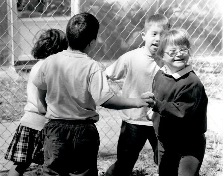 Four young children, three boys and one girl, playing and smiling near a chain-link fence in a park or schoolyard.