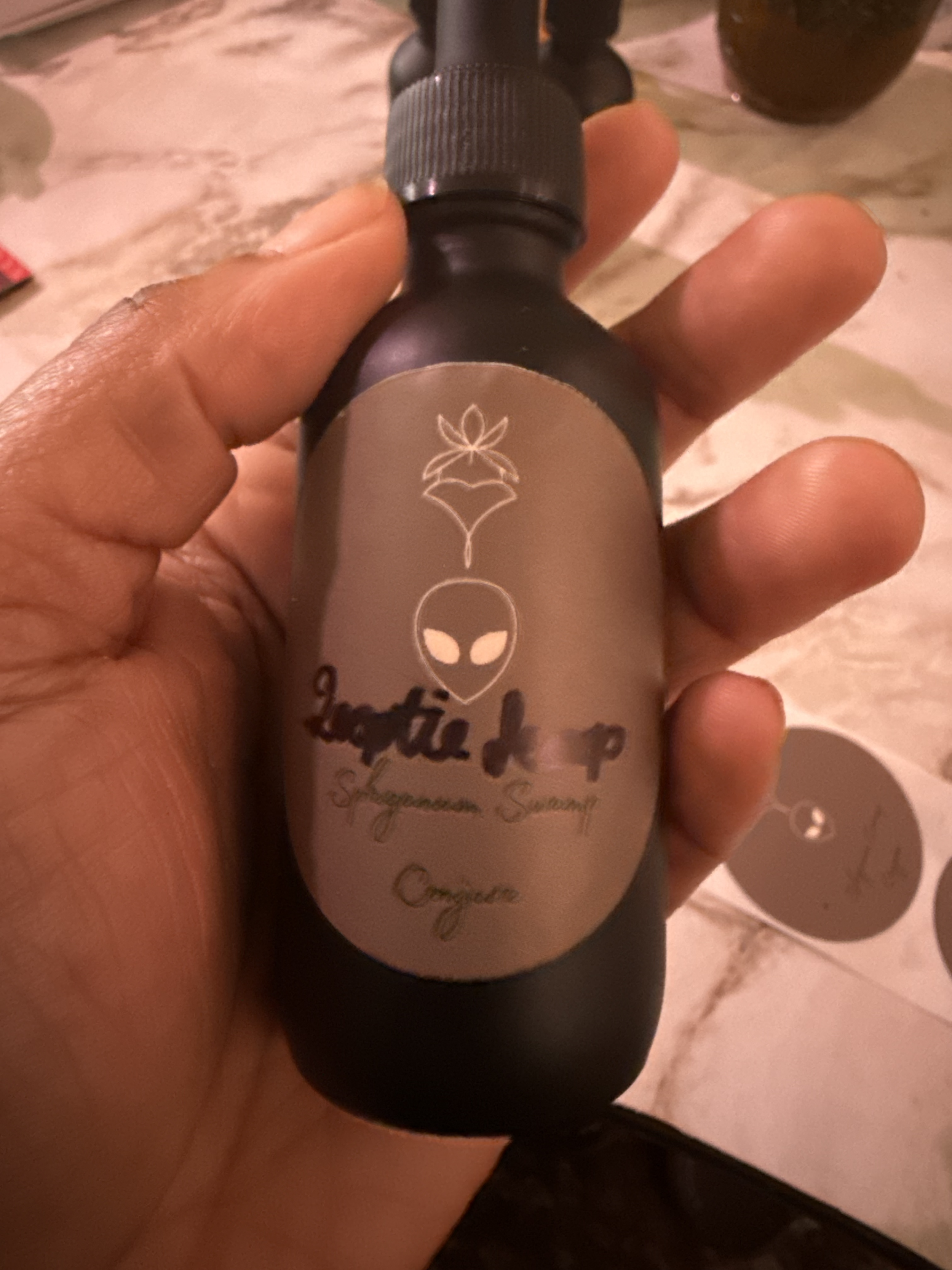 A hand holding a black spray bottle labeled 'Lustic Drop' with a logo of an alien face and a design of a plant or leaf.