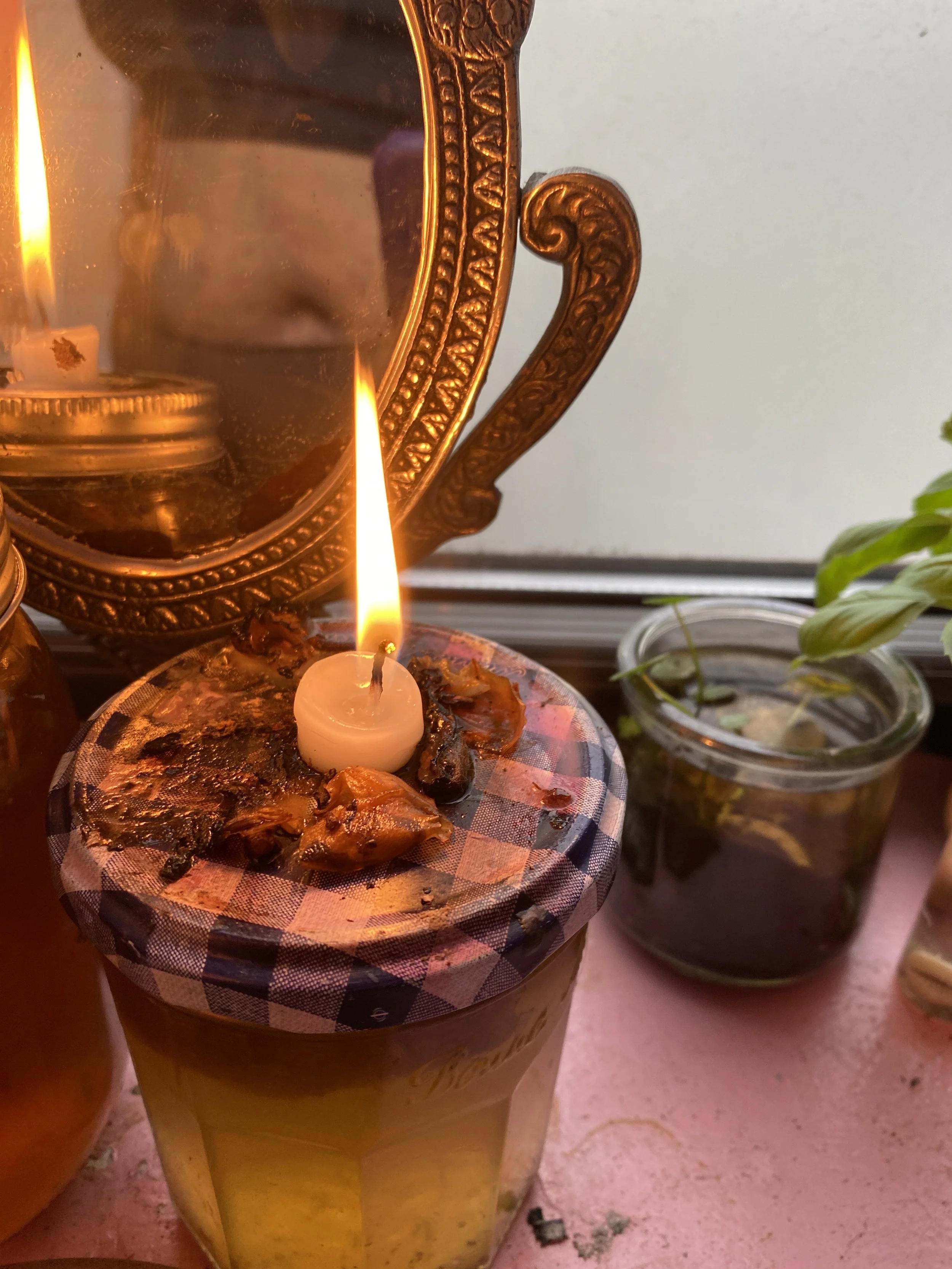 A lit birthday candle on a glass jar filled with honey or syrup, with a checkered cloth on top. In the background, a large ornate mirror with a golden frame and a small potted plant.