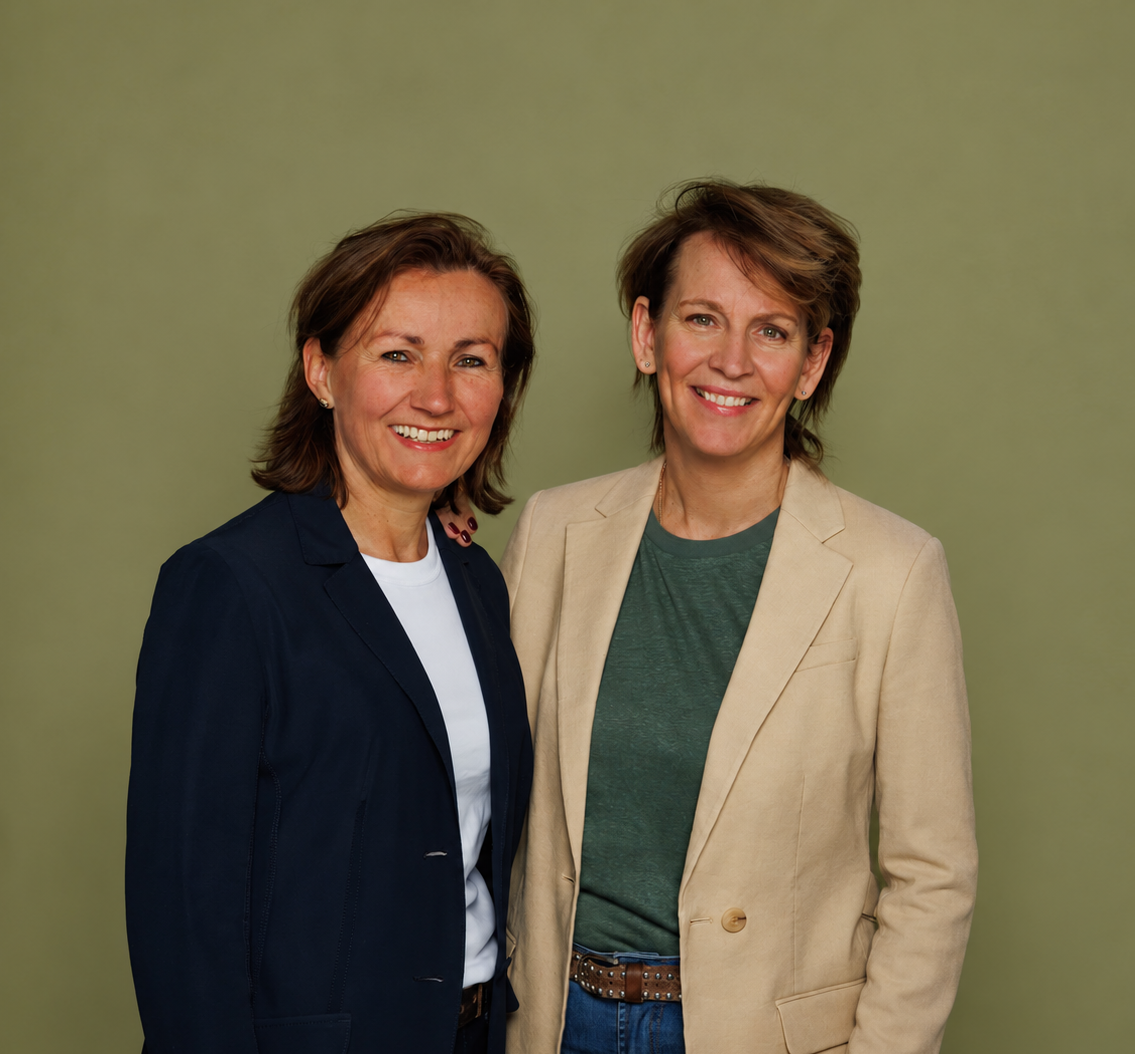 Twee vrouwen staan samen voor een lichtgroene achtergrond, glimlachend en gekleed in zakelijke casual kleding.