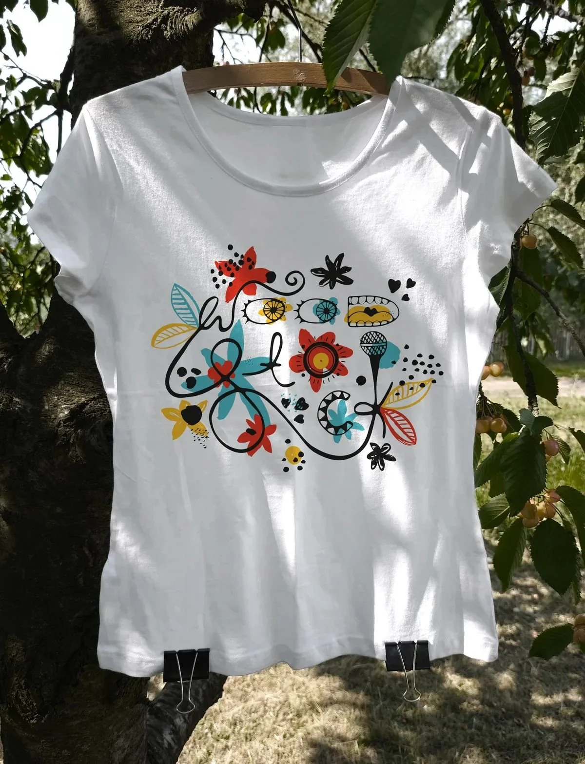 T-shirt with colorful flat-style event print hanging from a cherry tree branch in the garden — expressive composition of flowers, music, and summer energy.