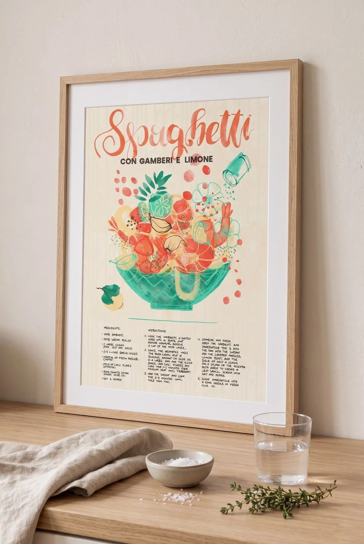 Framed print of an illustrated spaghetti recipe leaning against a kitchen wall, watercolor bowl of shrimp, lemon and garlic in coral and teal tones, styled on a wooden counter with a linen cloth, salt dish and glass of water.