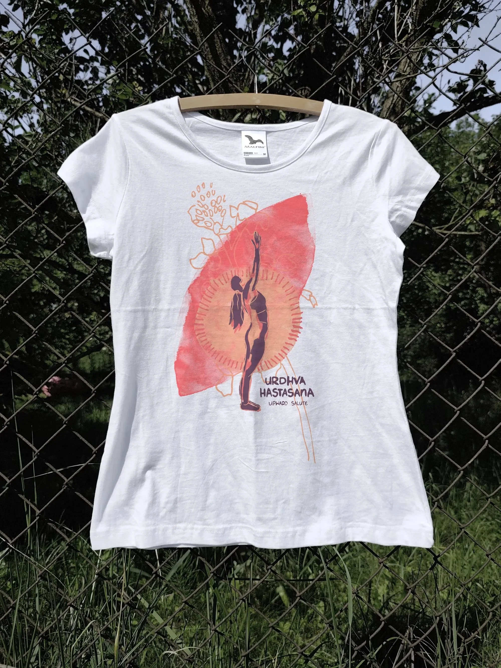 T-shirt featuring yoga pose Urdhva Hastasana from the Surya Namaskar series, photographed outdoors in a garden.