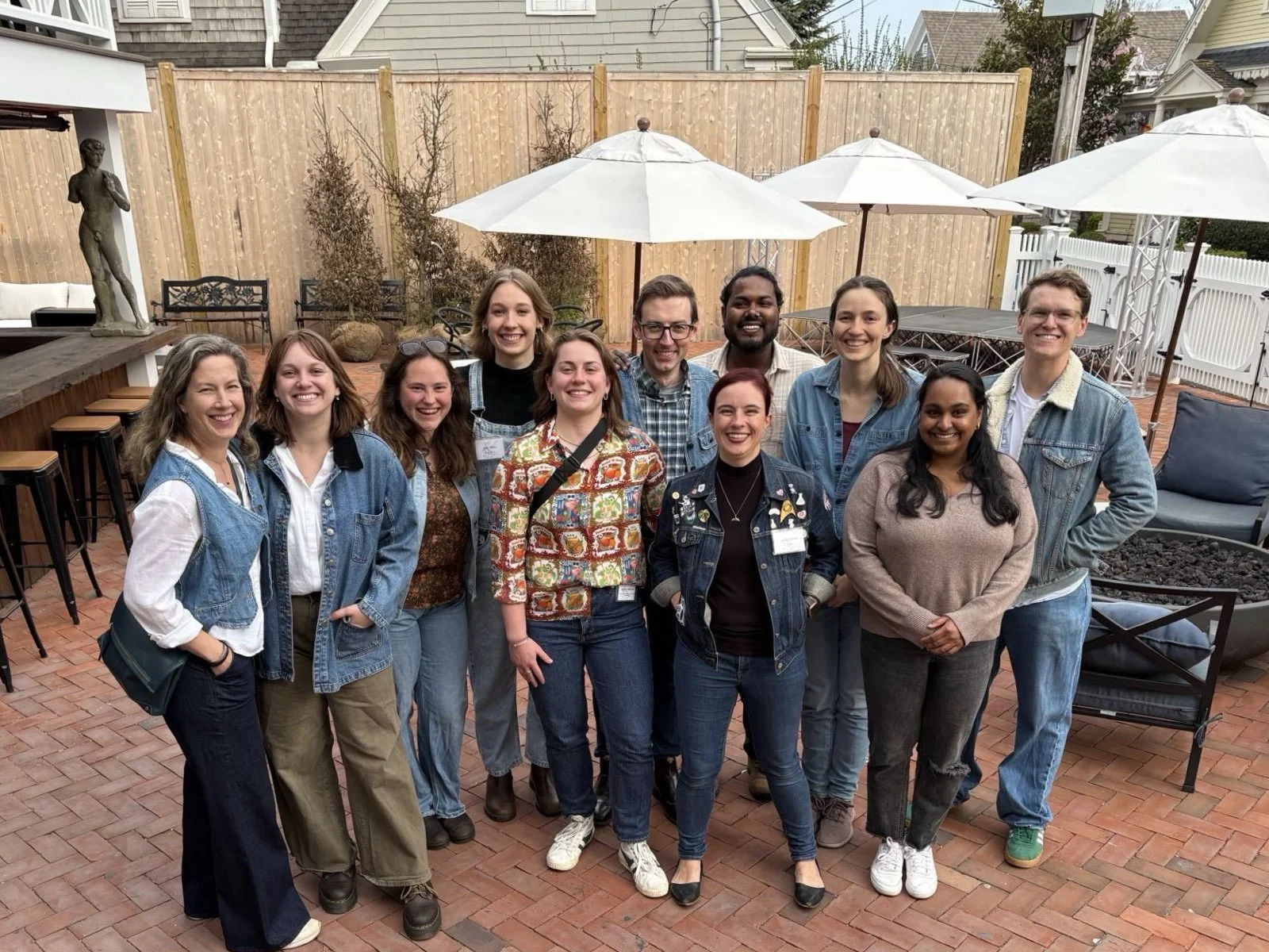 A group of twelve people, mostly women, standing closely together on a brick patio, smiling for the camera. The background includes patio umbrellas, outdoor furniture, a wooden fence, and some decorative plants.