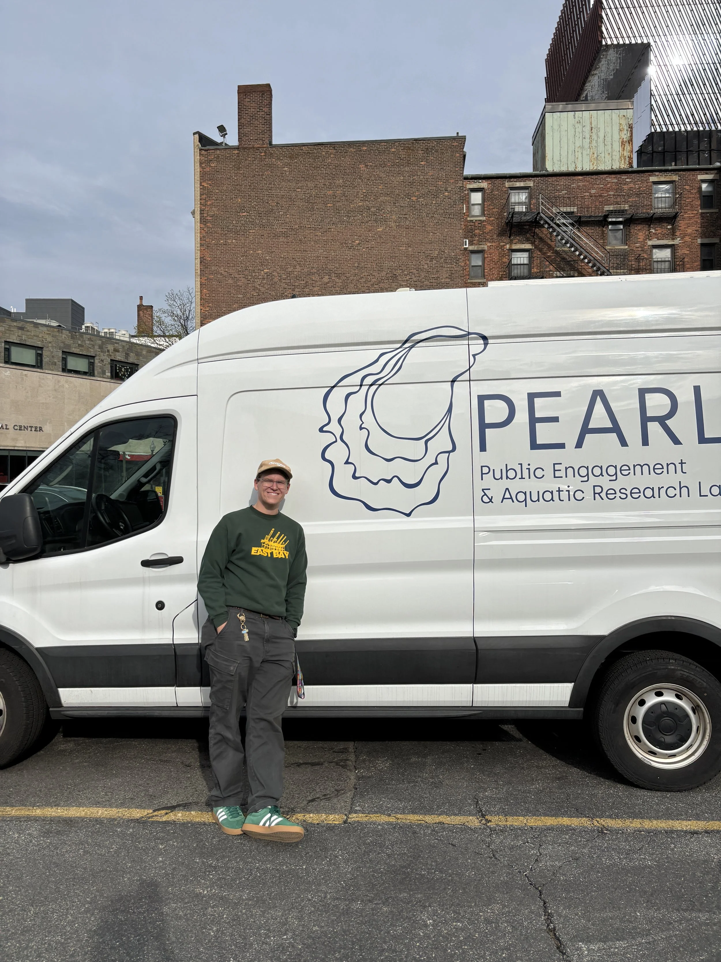 A man standing in front of a white van with the text 'PEARL Public Engagement & Aquatic Research Lab' and a seashell outline logo. The man is smiling, wearing a green sweatshirt, dark cargo pants, and green sneakers, and is standing on an asphalt parking lot with buildings in the background.