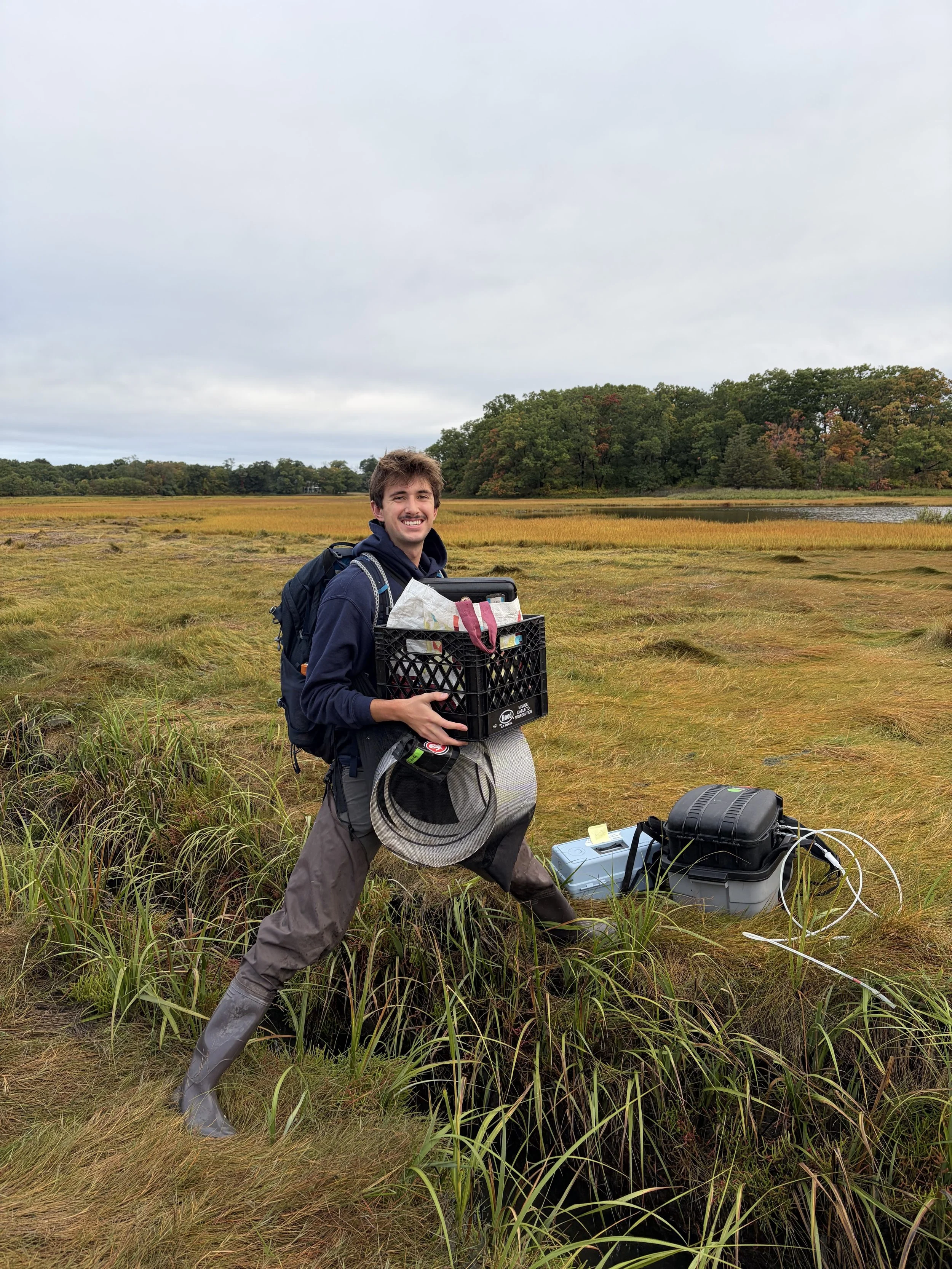 Young man outdoors in a marsh or wetland, dressed in waders and rain gear, holding equipment, with a happy expression and backpack, near some water and trees in the background.