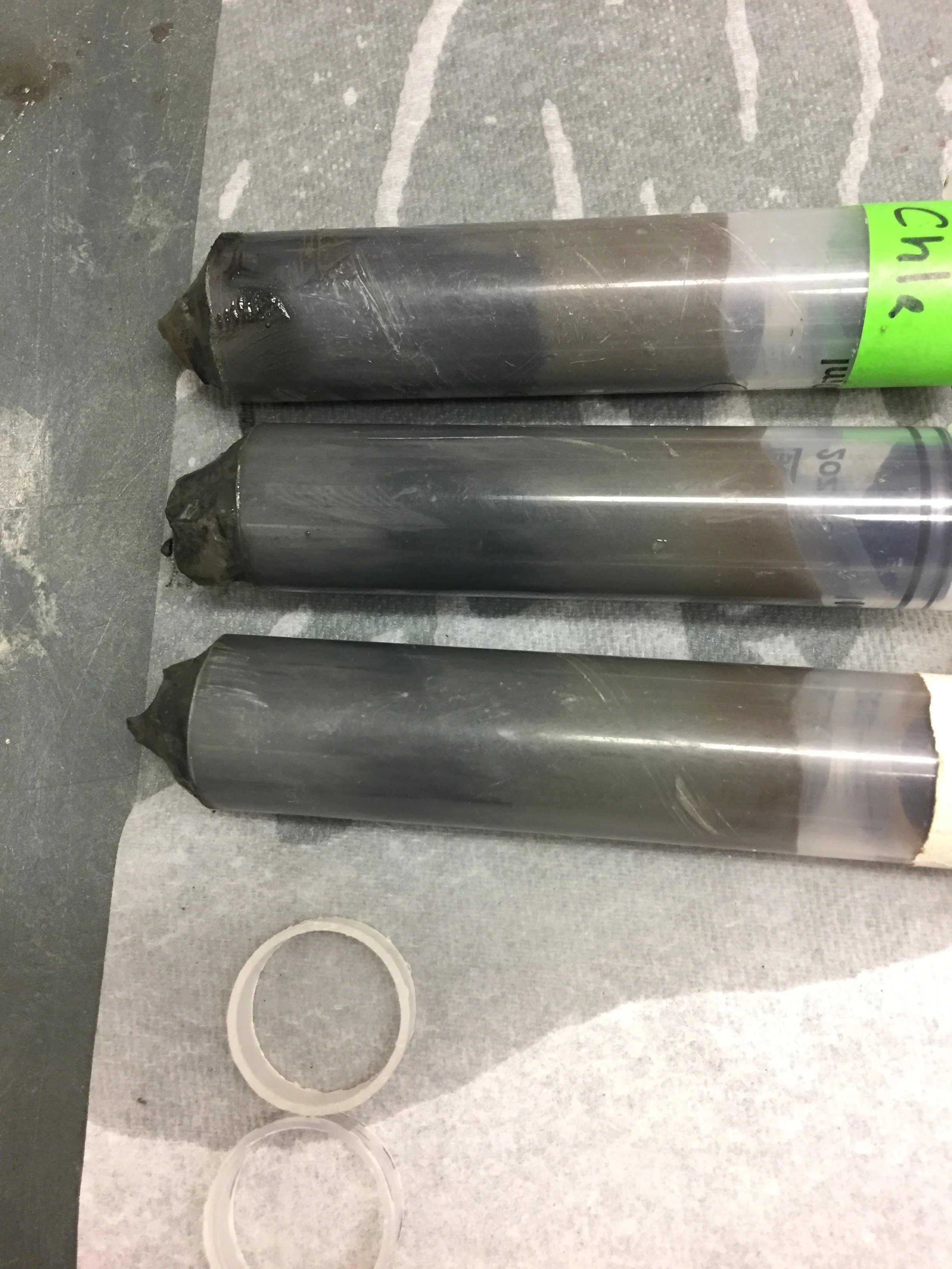 Three cylindrical metal drill bits with black tips, lying on a gray textured surface, with two transparent washers below them.