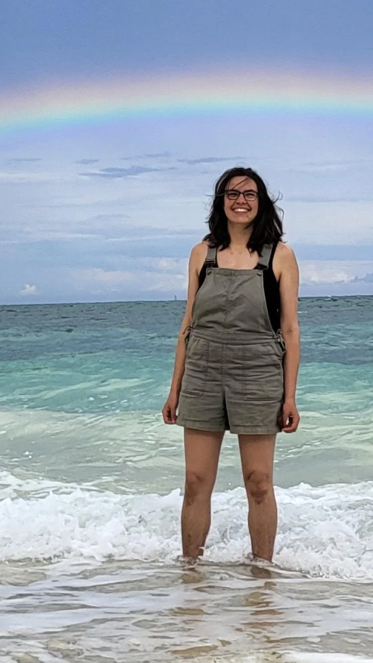 A young woman standing in the shallow ocean water at the beach with a rainbow visible in the sky above her.