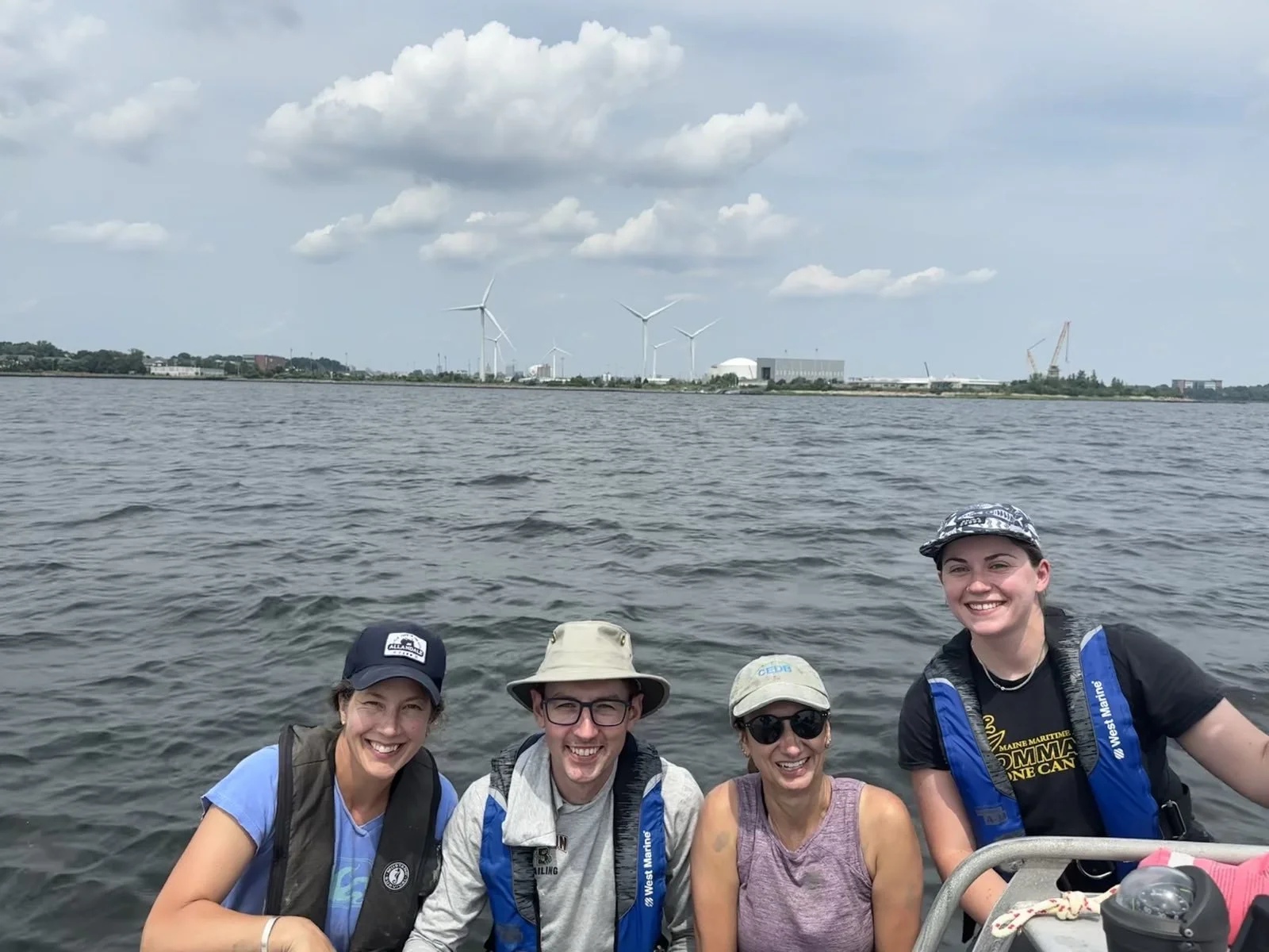 Four smiling people on a boat, with water and a shoreline with wind turbines and industrial buildings in the background.