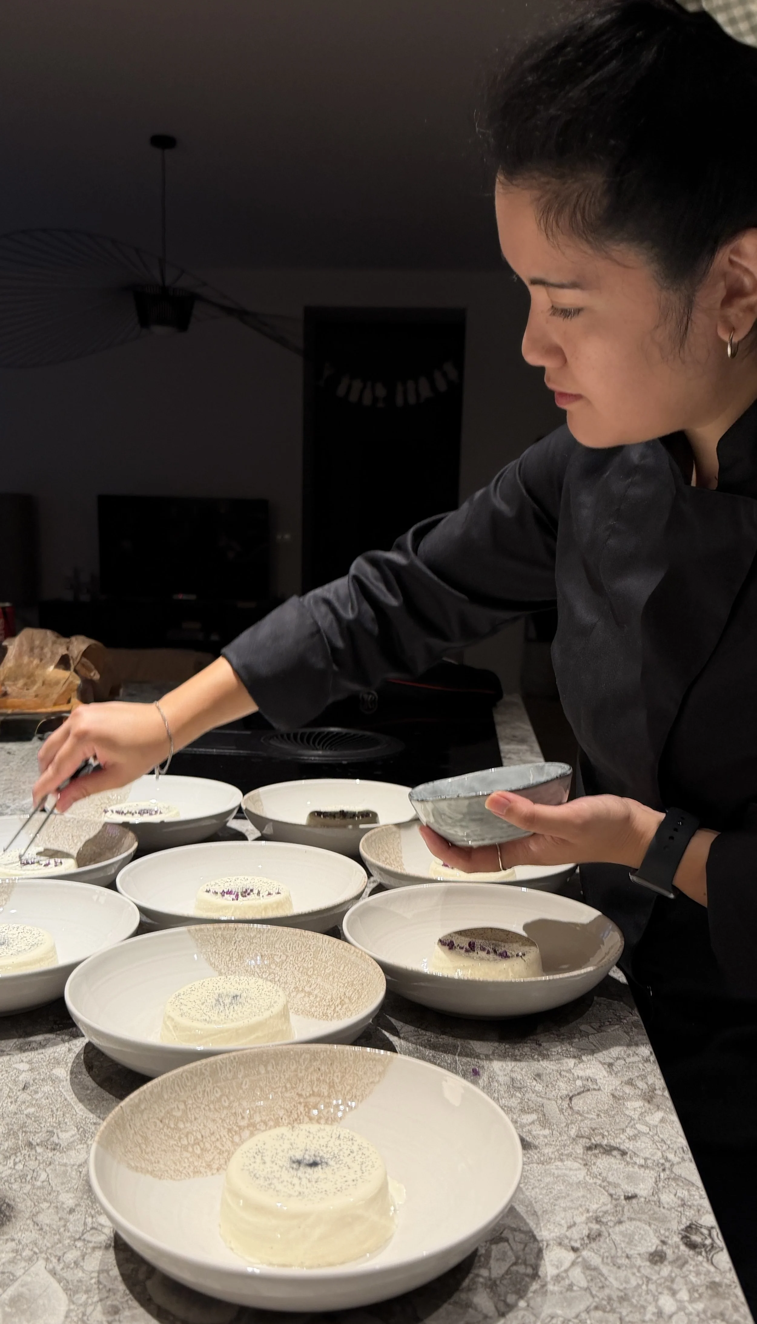 Une femme en cuisine, portant un uniforme noir, met en place un plat dans une cuisine moderne avec plusieurs plats blancs garnis de nourriture et de décoration, tels que de la mousse ou du gâteau.
