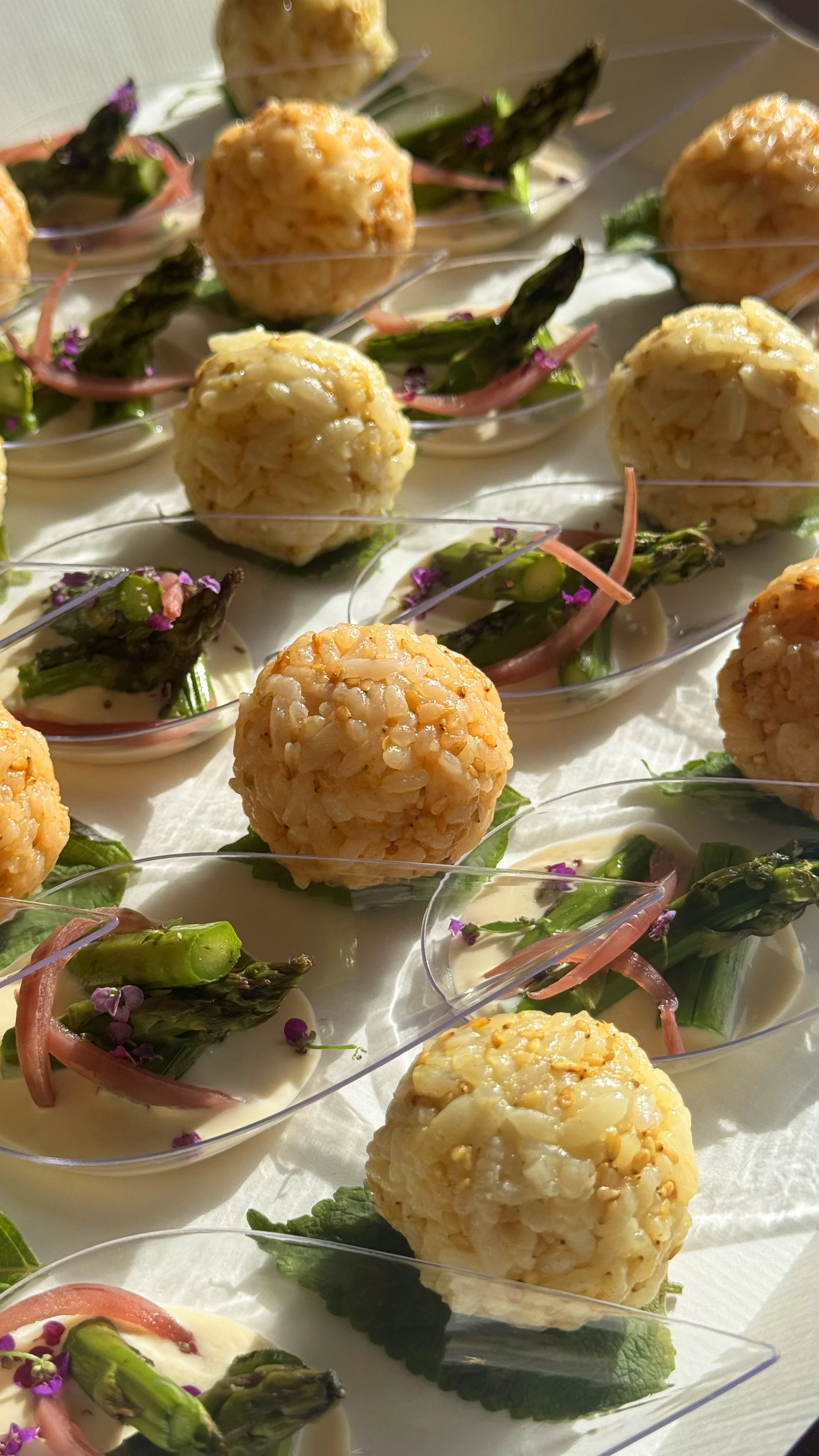 Bouchées apéritives avec des boulettes de riz, accompagnées d'asperges et de petites fleurs violettes, servies dans des coupelles en plastique transparentes pour une prestation de traiteur événementiel en Bourgogne