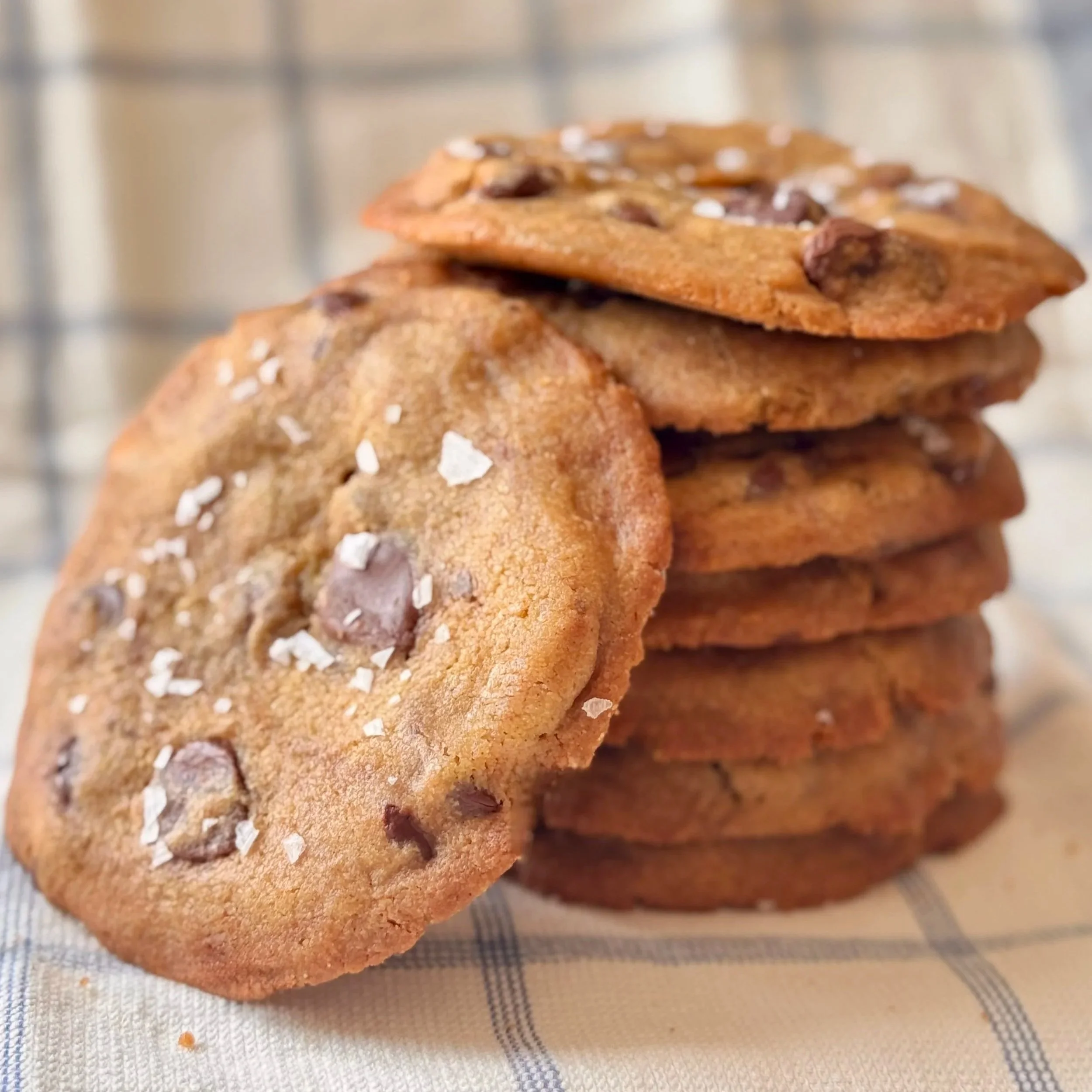 A stack of tahini chocolate chip cookies sprinkled with sea salt. One cookie leans against the side of the stack, all on a checkered cloth.