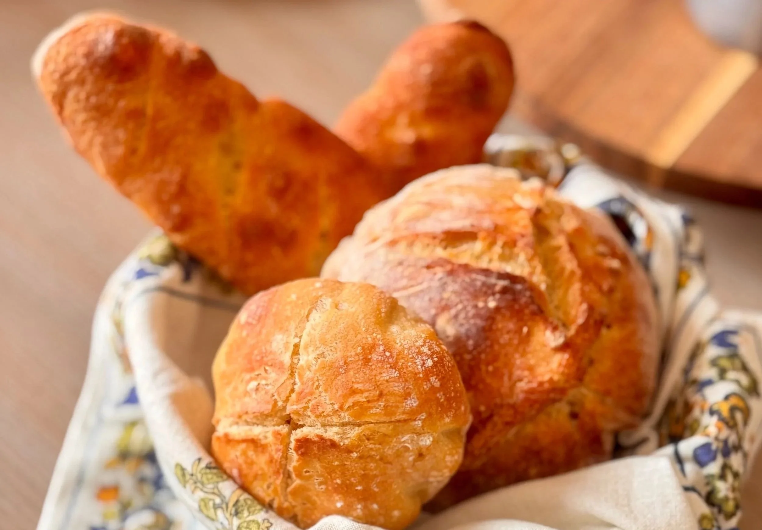 A basket with three freshly baked bread rolls, one big and two small, placed on a decorative cloth, with a wooden cutting board in the background.