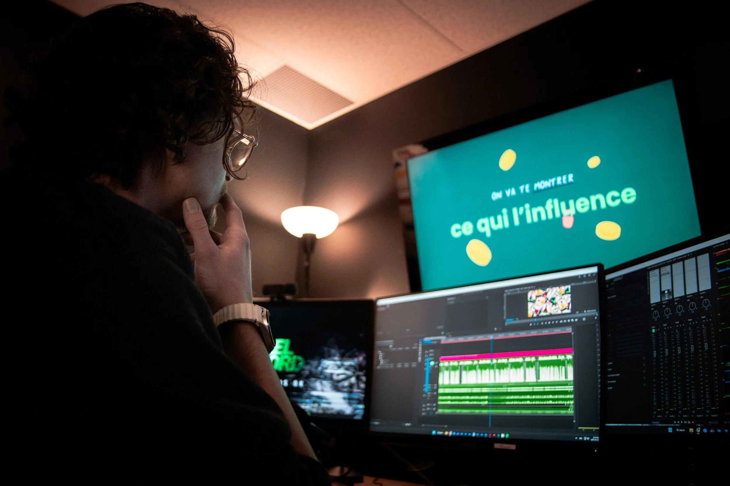 Une personne travaillant sur un montage vidéo dans une pièce sombre, avec un grand écran affichant un message en français.