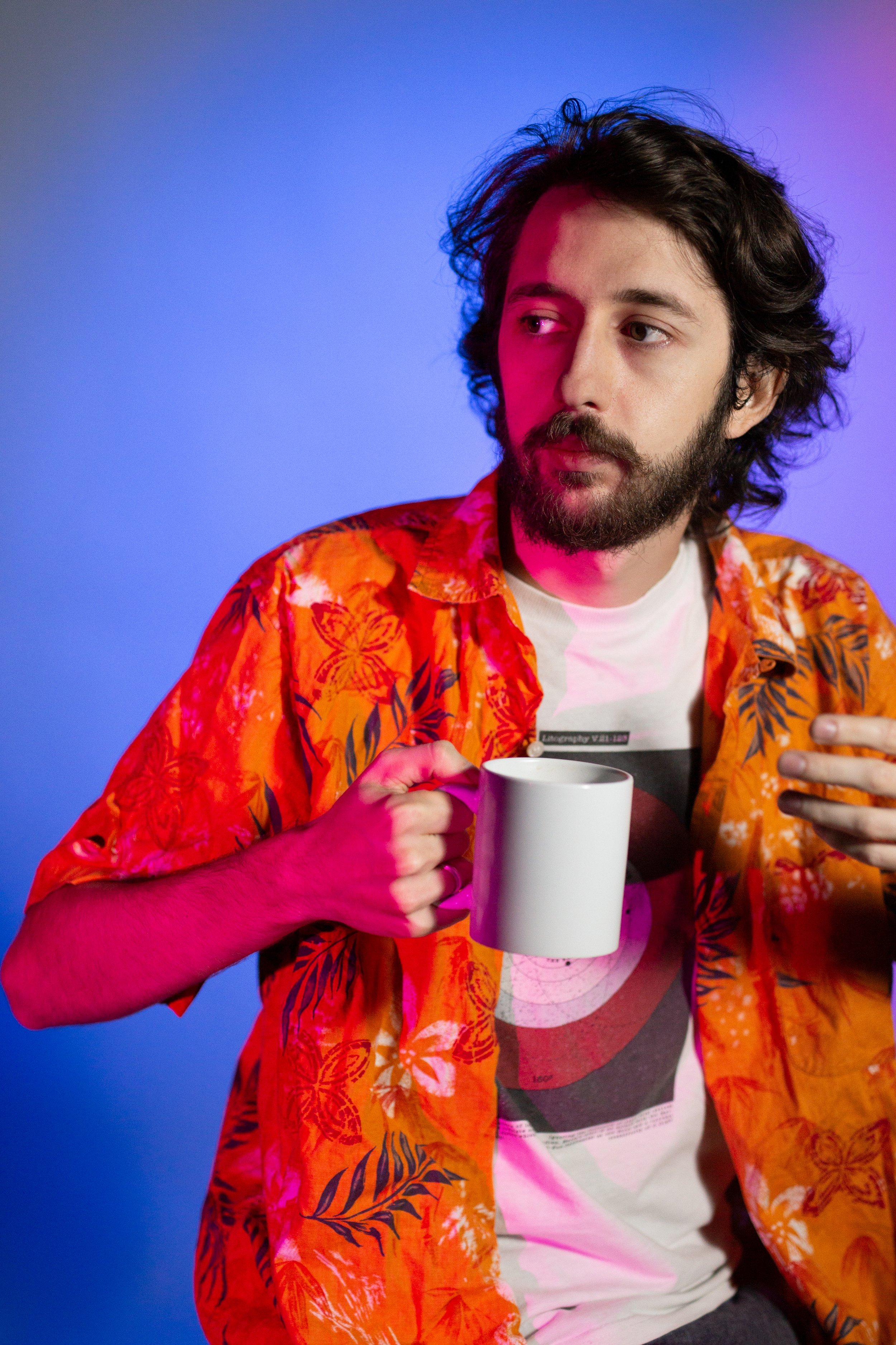Homme aux cheveux longs, barbe, portant une chemise hawaiienne orange avec motifs floraux, tient une tasse blanche, regarde sur le côté, fond violet et bleu lumineux.