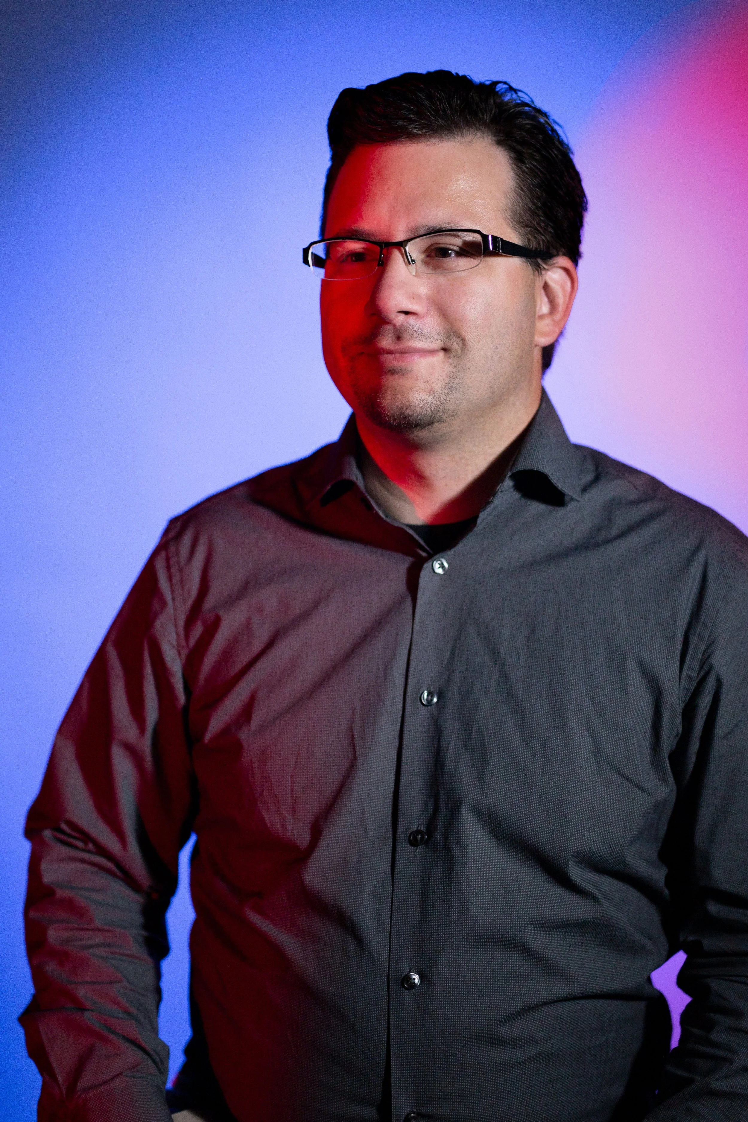 Un homme portant une chemise noire et des lunettes, souriant légèrement, avec un fond coloré dégradé bleu et rose.
