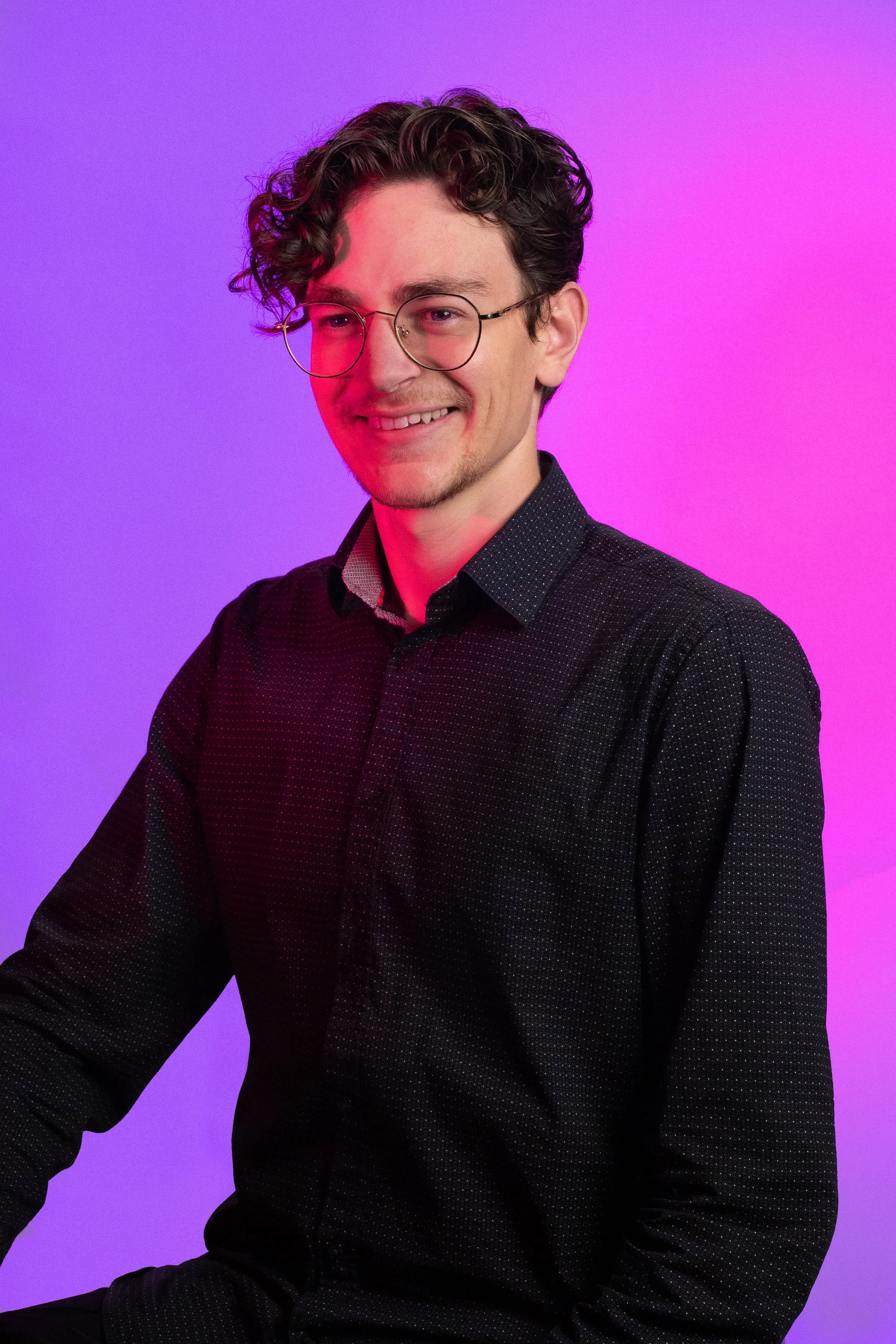 Jeune homme avec lunettes, souriant, debout devant un fond violet et rose.