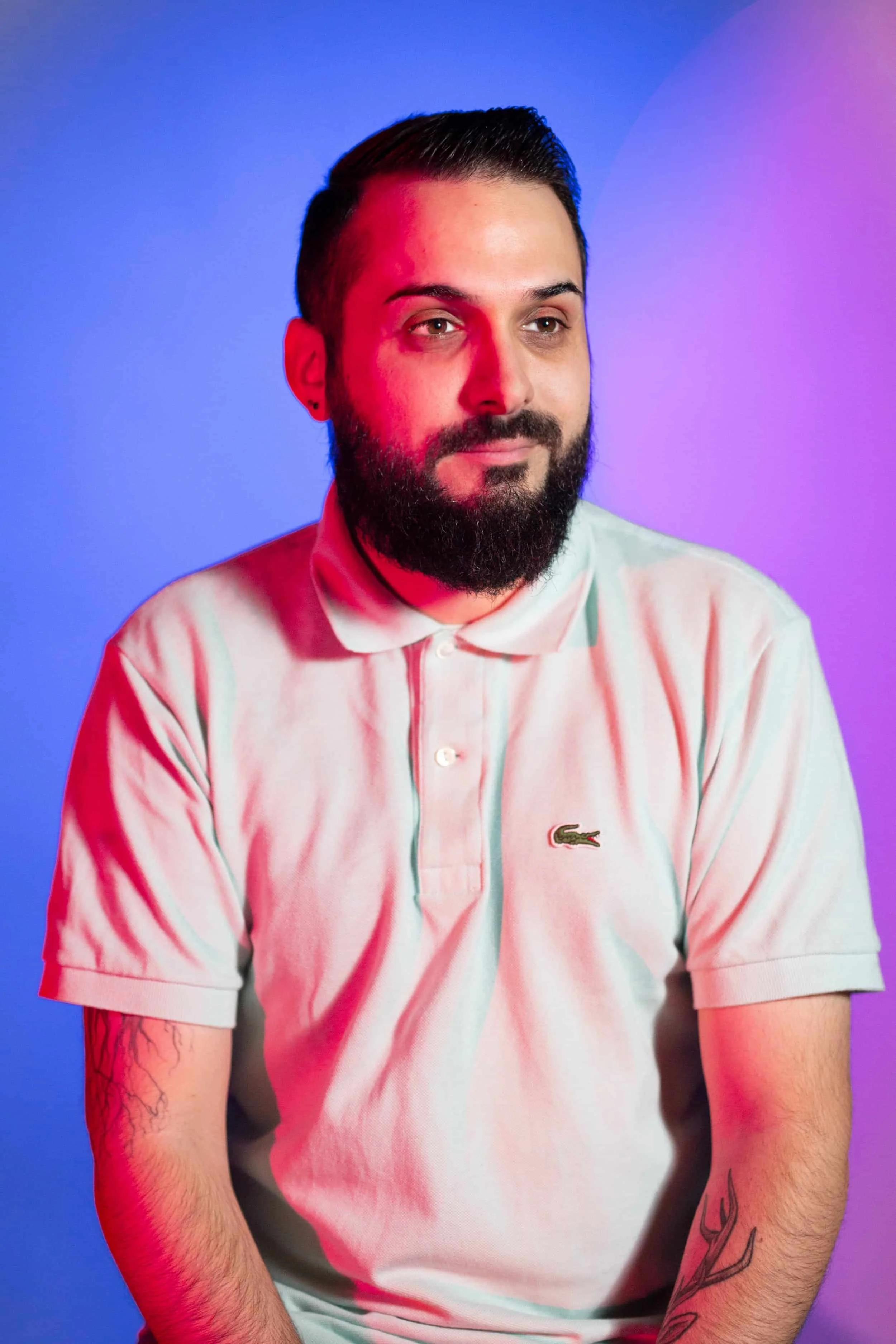 Réalisateur / Ressources Humaines. Portrait d'un homme avec moustache, barbe, portant un polo blanc avec un logo Lacoste, devant un fond à dégradé de couleurs (bleu et violet).