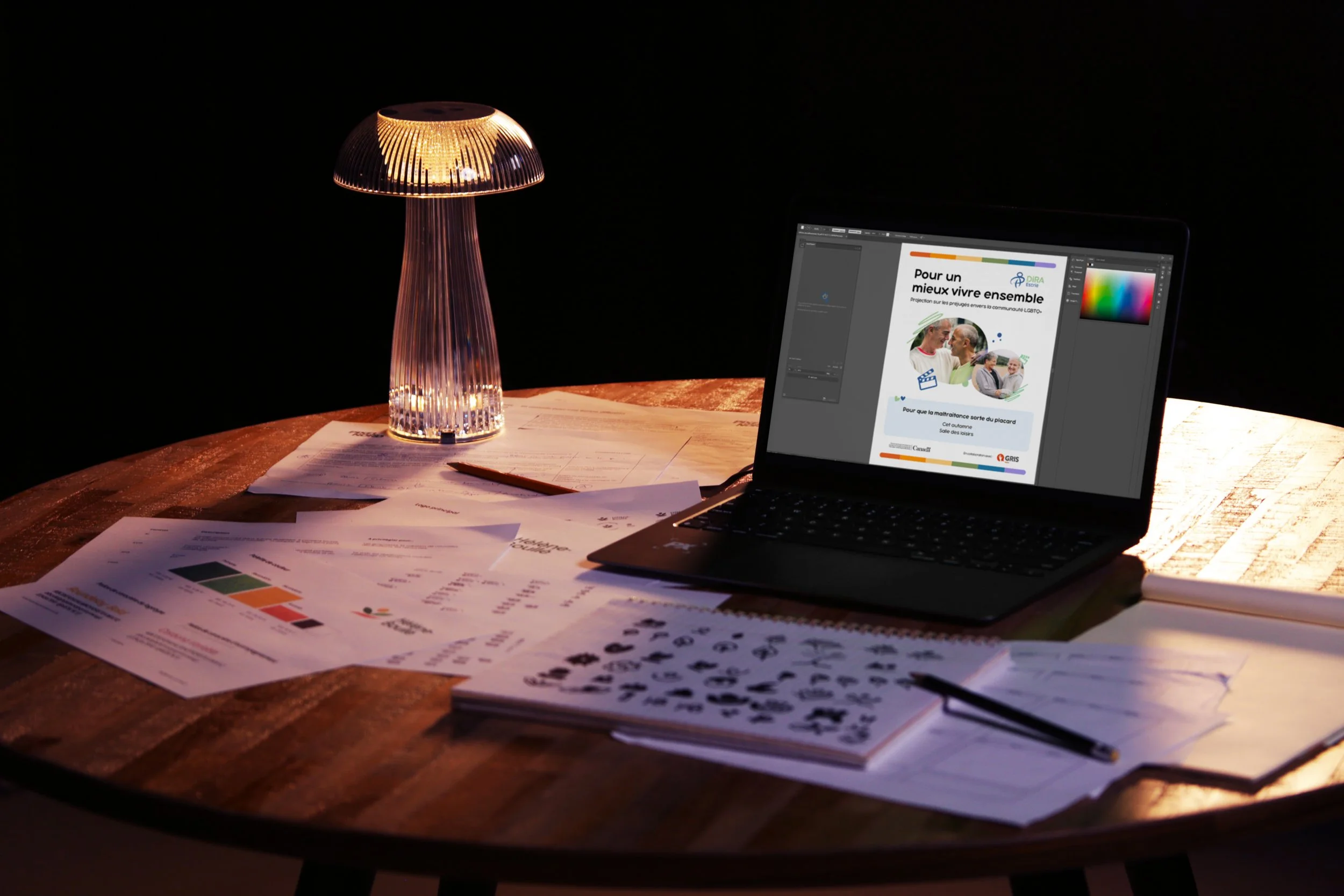 Table de travail avec des papiers, un livre d'images, un ordinateur portable affichant un document en français, une lampe moderne allumée et un crayon.