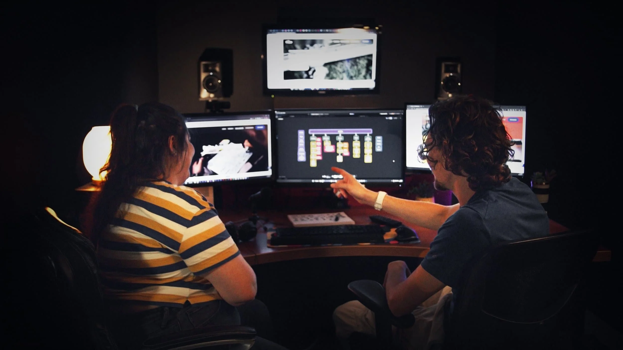 Deux personnes travaillent dans un studio de montage vidéo avec trois écrans derrière elles, affichant des logiciels de montage et des images.