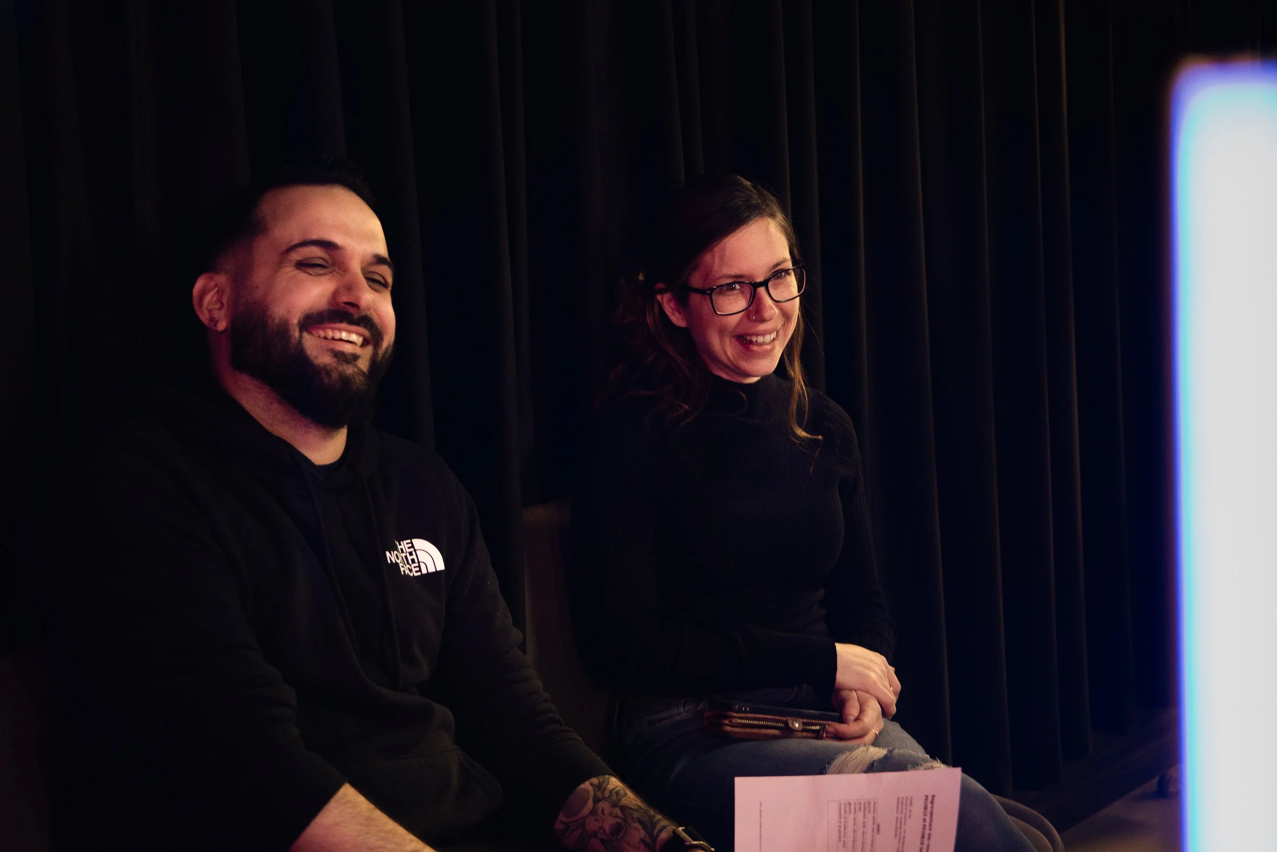 Deux personnes assises côte à côte, rie et sourient, en face d'un mur noir avec éclairage doux.