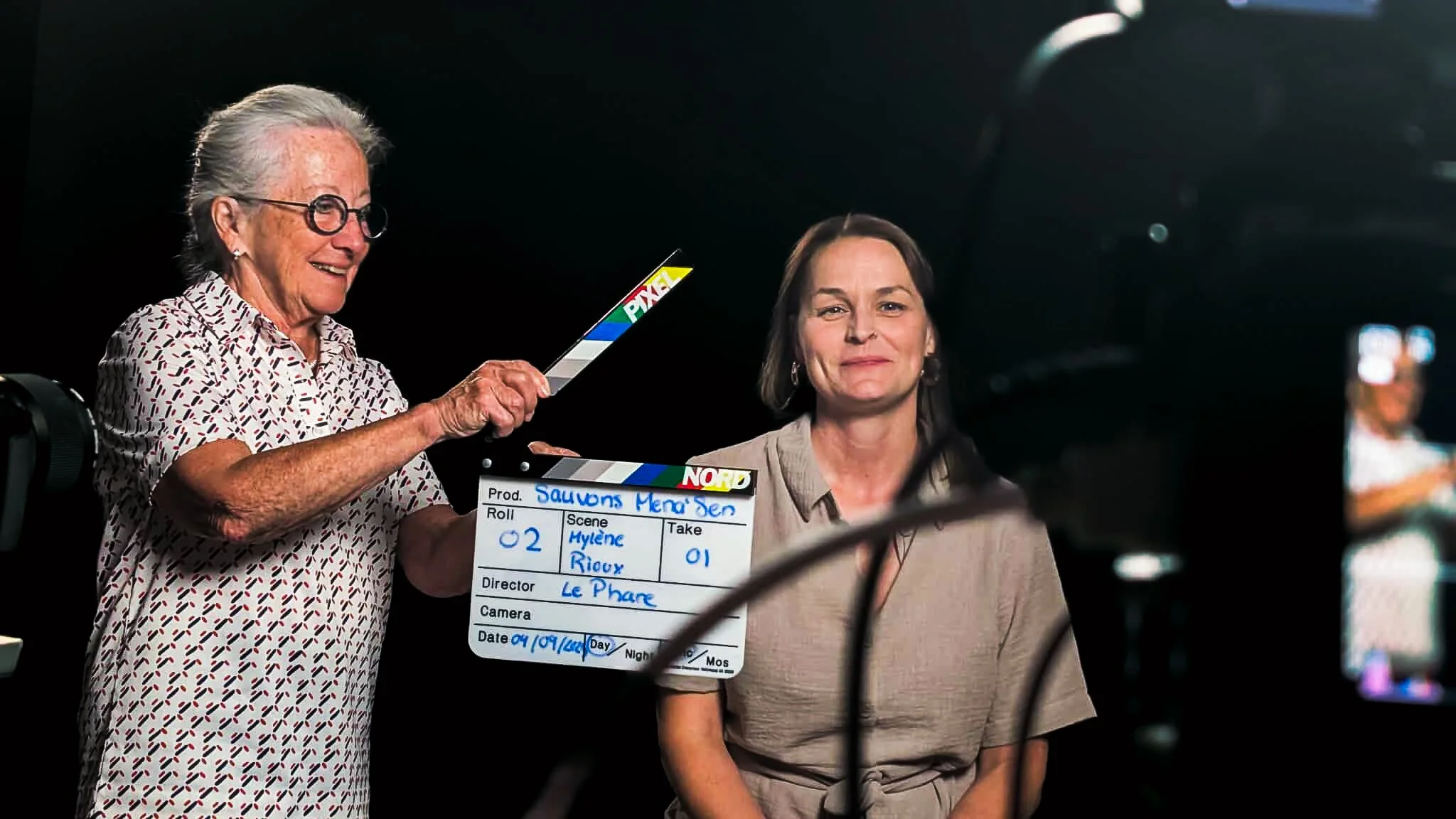 Deux femmes participant à une scène de tournage, une tient une clap de film et l'autre est assise devant un fond noir, preuve de l'atmosphère d'un plateau de film ou de télévision.