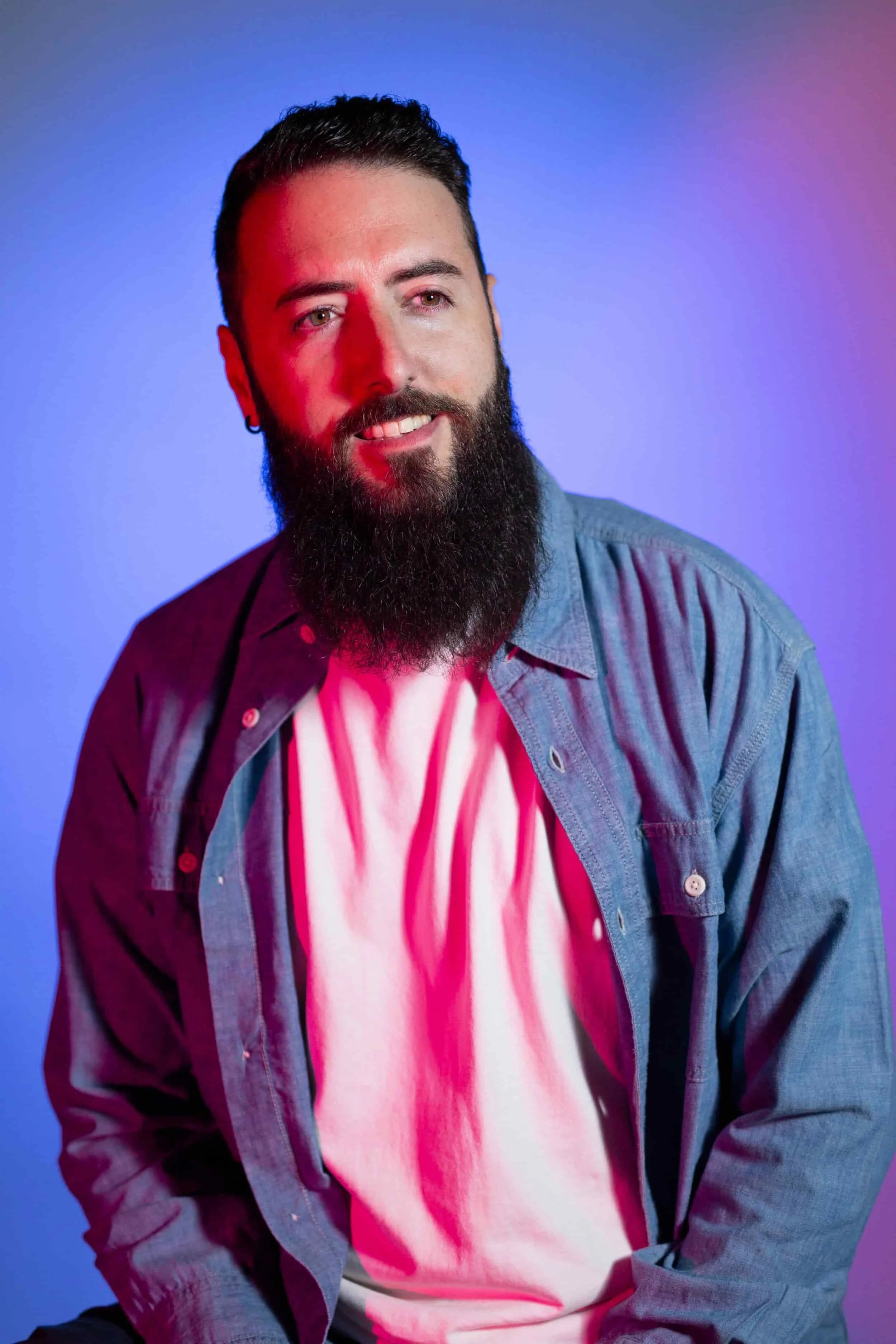 Réalisateur / Directeur Photo. Un homme avec une barbe, portant une chemise en jean et un t-shirt blanc, est en train de sourire. Il y a un fond coloré en dégradé de bleu et rose.