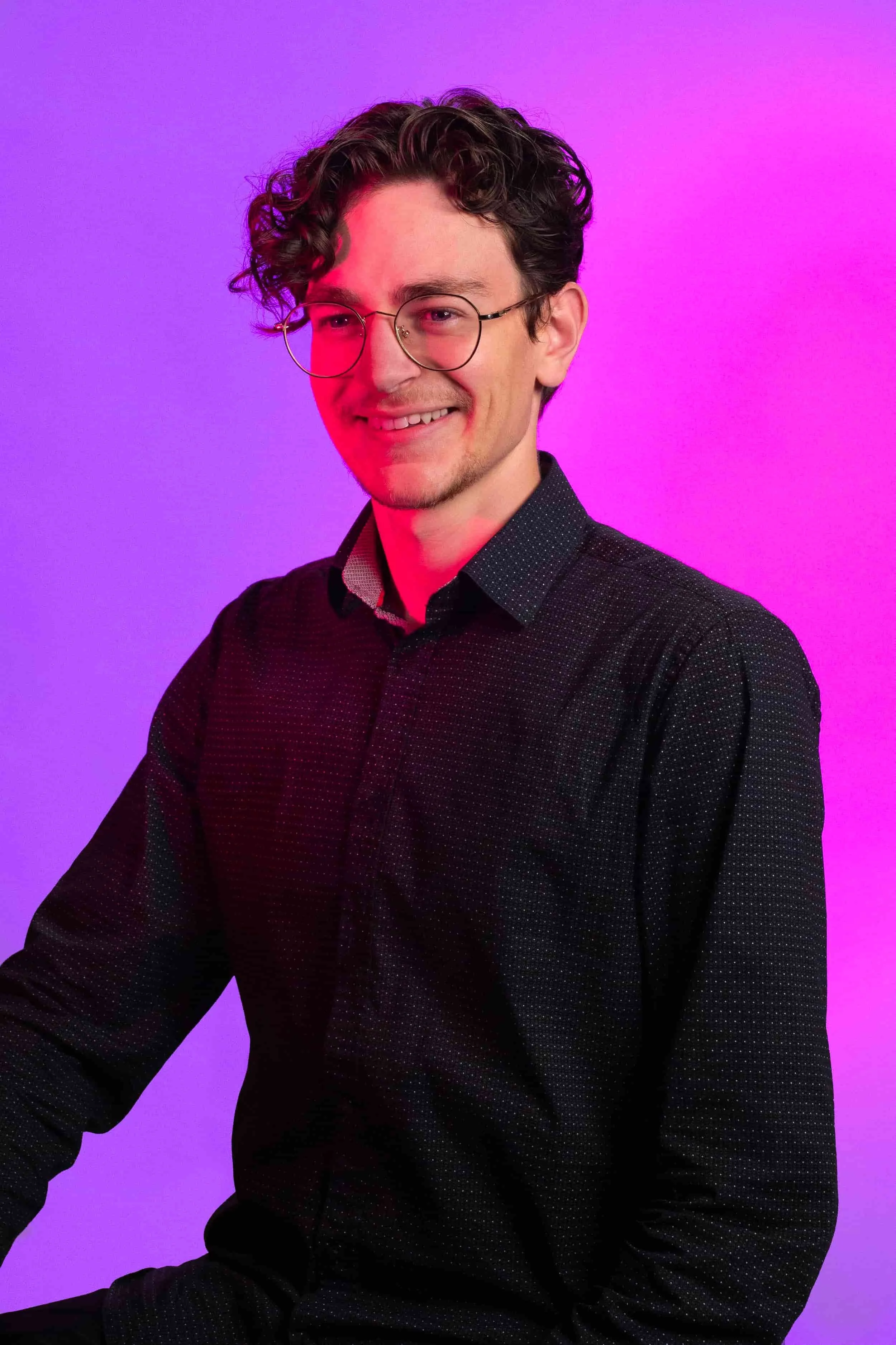 Monteur / Motion Designer. Jeune homme avec lunettes, souriant, debout devant un fond violet et rose.