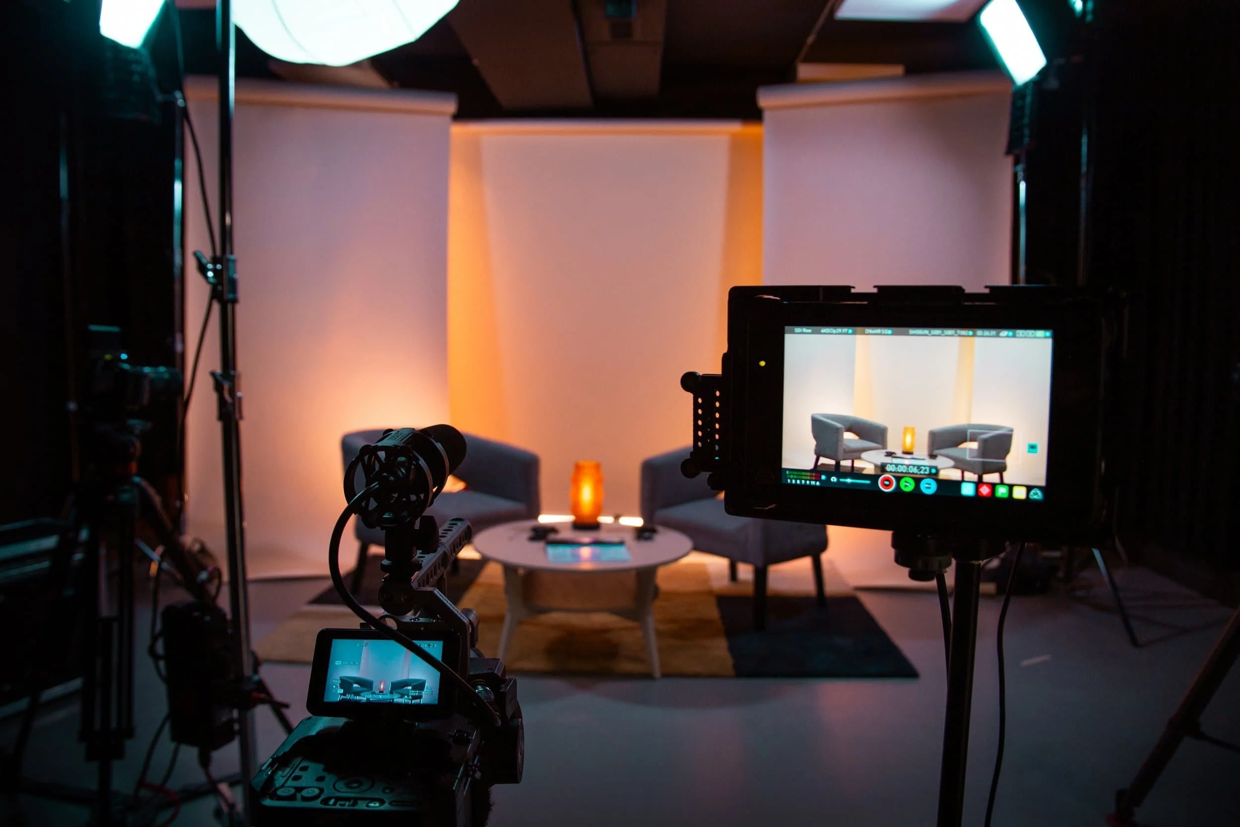Studio d'enregistrement avec deux fauteuils gris, une table basse ronde, un luminaire orange, et un fond blanc. Présence d'une caméra et d'éclairage pour la mise en scène.