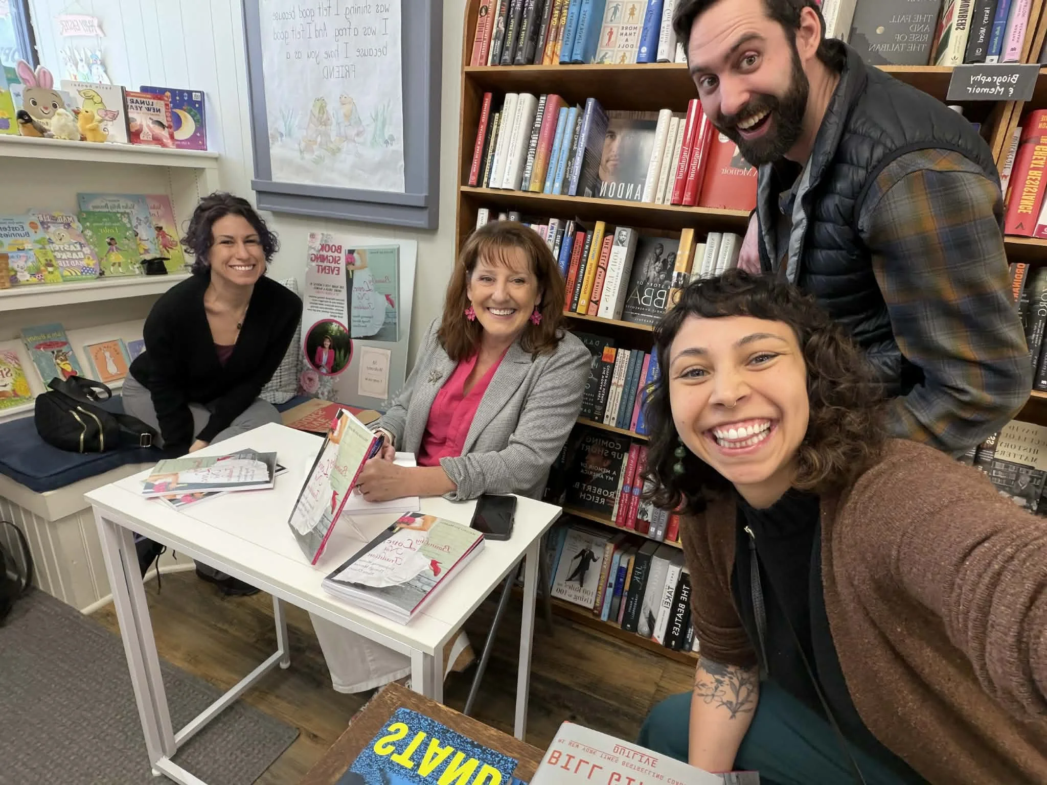 Four people smiling and taking a selfie in a bookstore. Two women are seated at a table with books in front of them, and a man and a woman are standing behind them. Bookshelves filled with books are visible in the background.