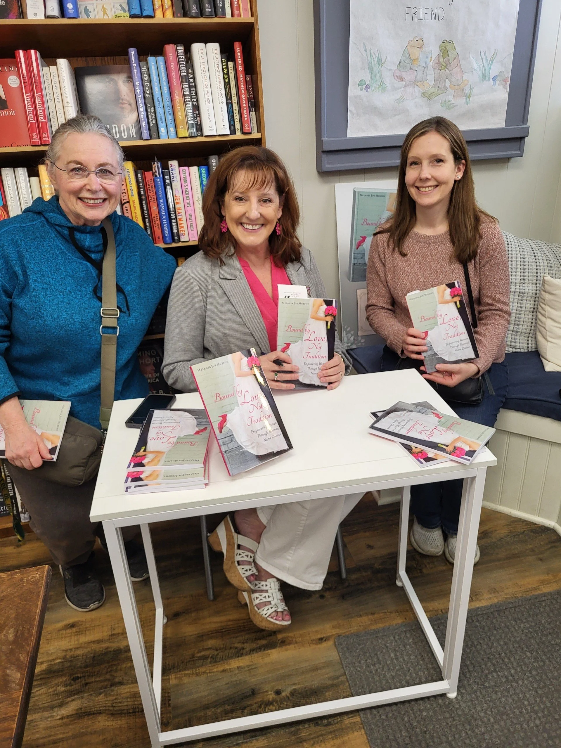 Three women sitting and standing around a white table in a bookstore, holding copies of a book titled 'Bound by Love Not Tradition.' The woman on the left is wearing a blue hoodie, the woman in the middle is wearing a grey blazer, and the woman on the right is wearing a pinkish sweater. There are additional books and promotional materials on the table, with bookshelves and a drawing of frogs in the background.