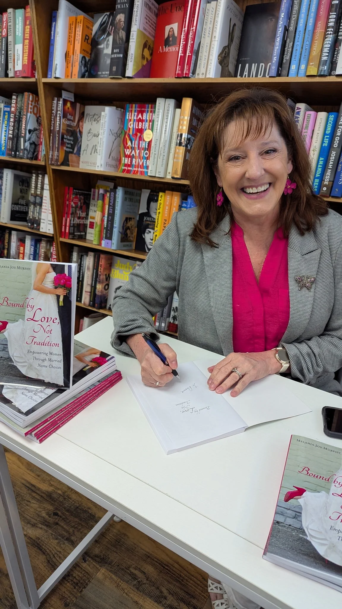 A woman with brown hair and pink earrings, wearing a gray blazer and hot pink blouse, sitting at a table in front of a bookshelf, signing a book during a book signing event.