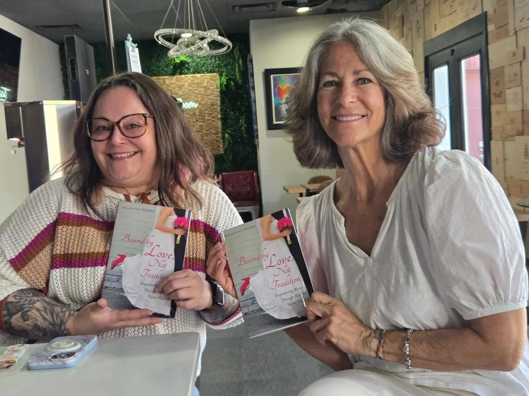 Two women smiling and holding books titled 'Bound by Love, Not Tradition' in a cozy, well-lit room with modern decor.