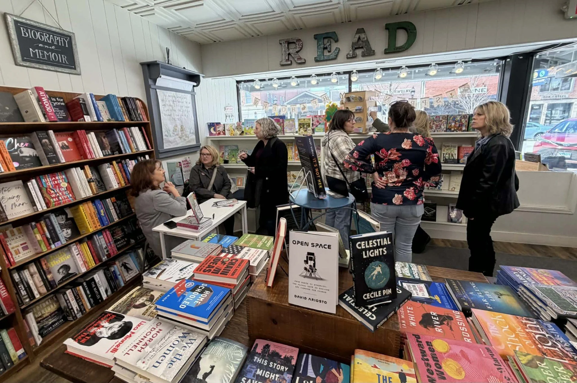 Inside a bookstore with shelves filled with books, several women are engaged in conversation near the front window, which displays decorative letters spelling 'READ'. A sign on a table reads 'Open Space' and 'Celestial Lights' by Cécile Pin. Customers browse and discuss books in a cozy, well-lit environment.