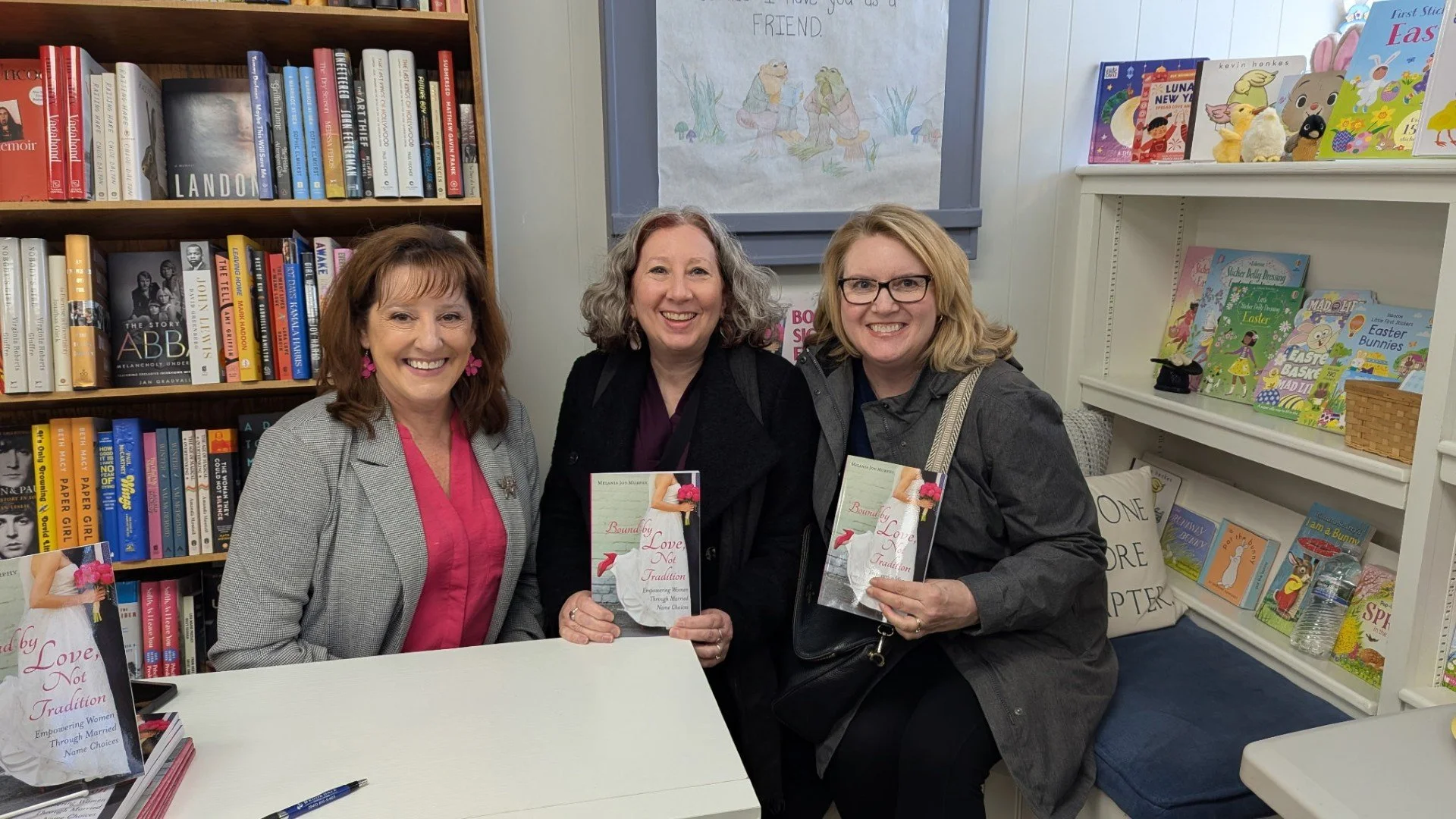 Three women sitting at a table in a bookstore, holding copies of a book titled 'Bound by Love, Not Tradition'. Bookshelves filled with children's books and novels are visible behind them.